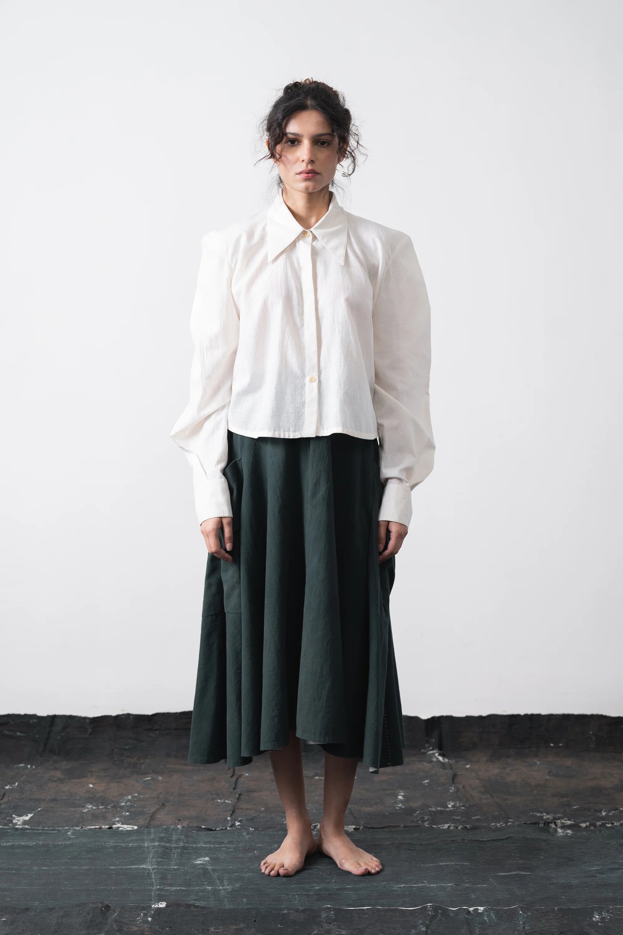 A model stands barefoot against a plain white wall, wearing a white long-sleeve button-up shirt with a high collar and a midi skirt.