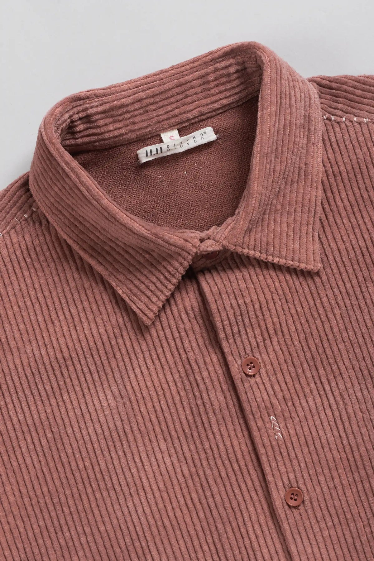 Close-up of a ribbed corduroy button-up shirt in a rough pink or mauve color, showing the collar, buttons, and inner neck label.