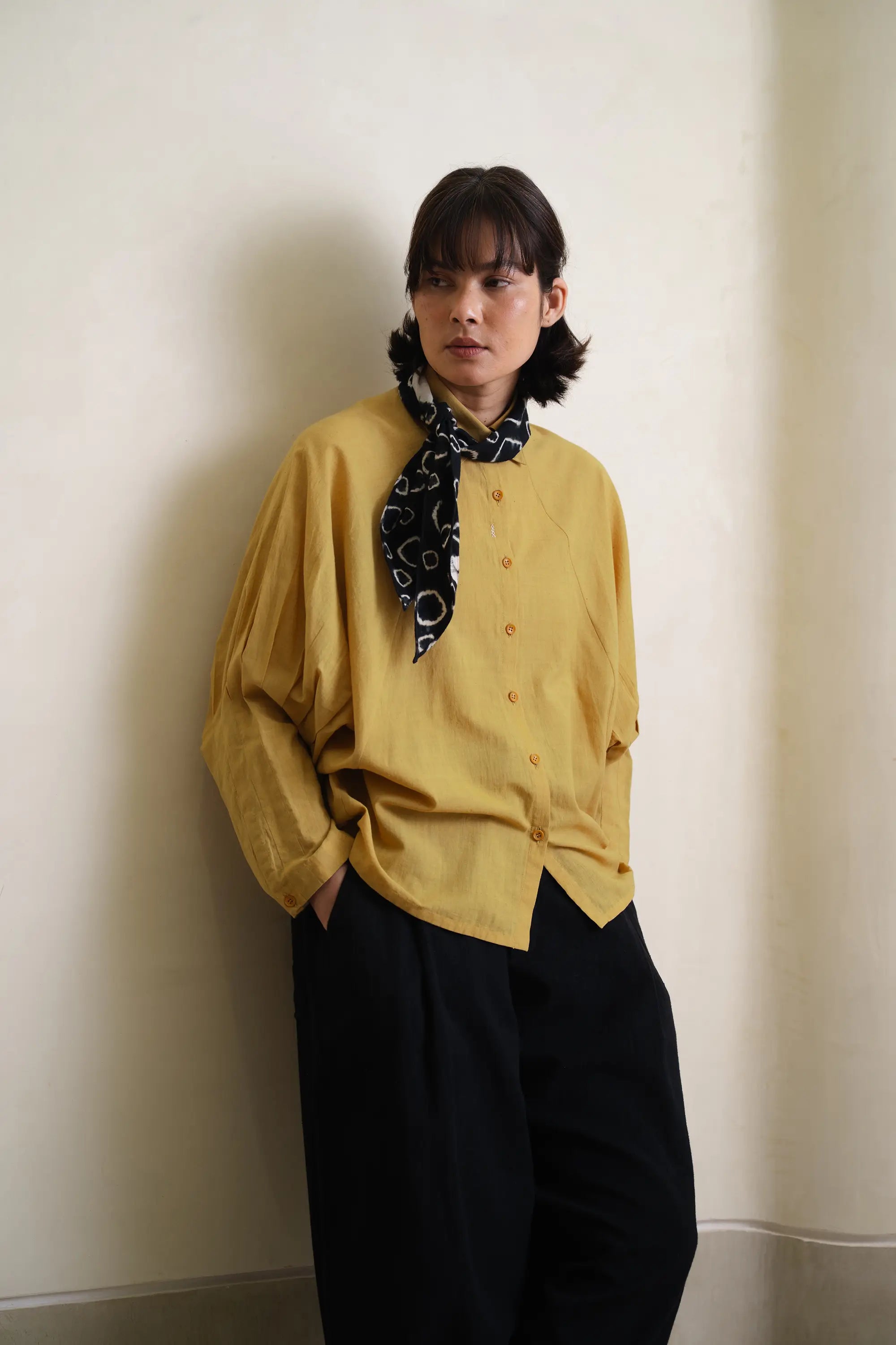 Person wearing a yellow shirt with a black patterned scarf, standing against a plain wall.