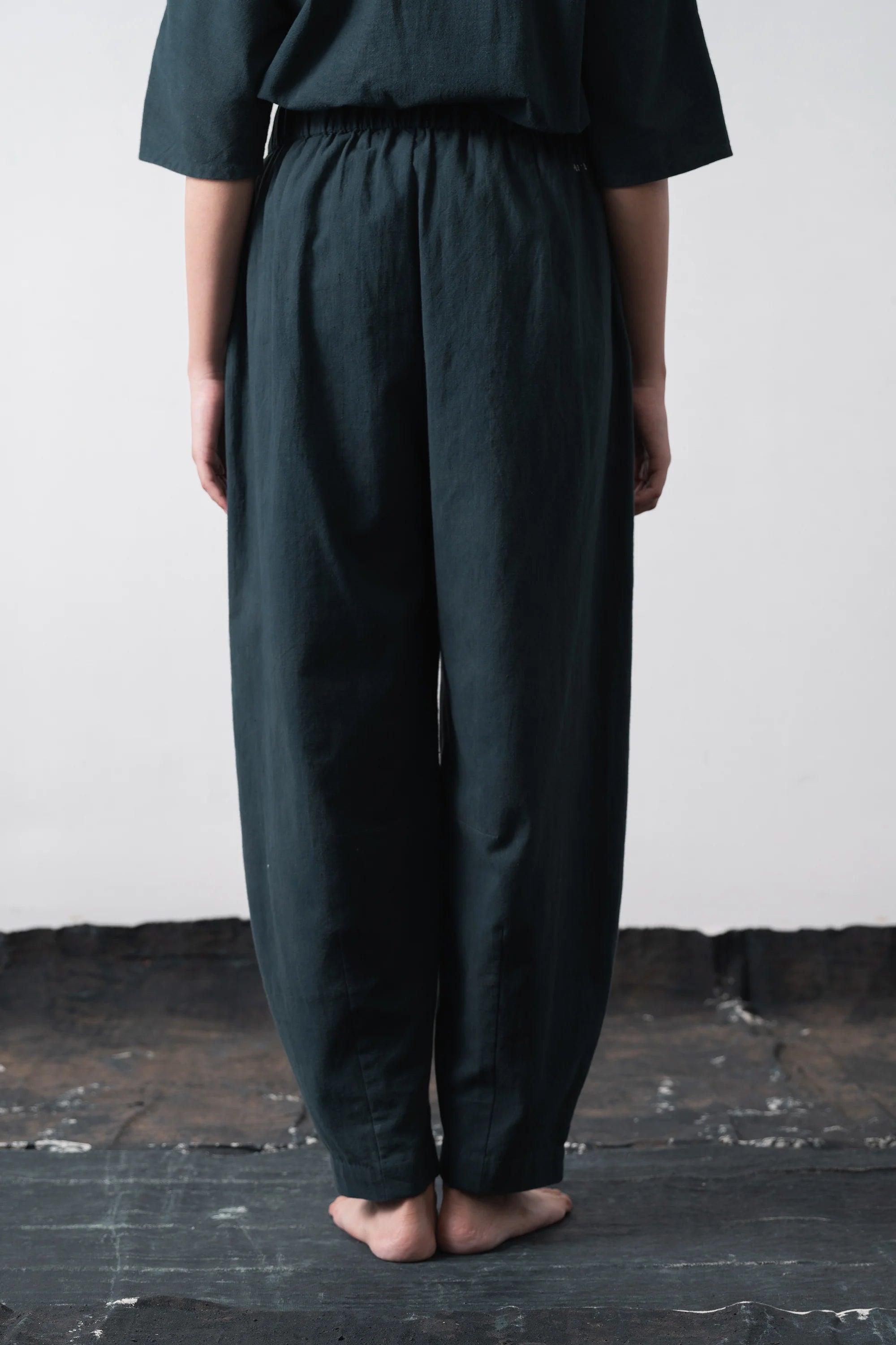 A person viewed from behind wearing loose, black indigo wide-leg trousers with an elastic waistband and side pockets, standing barefoot against a grey wall and dark floor.