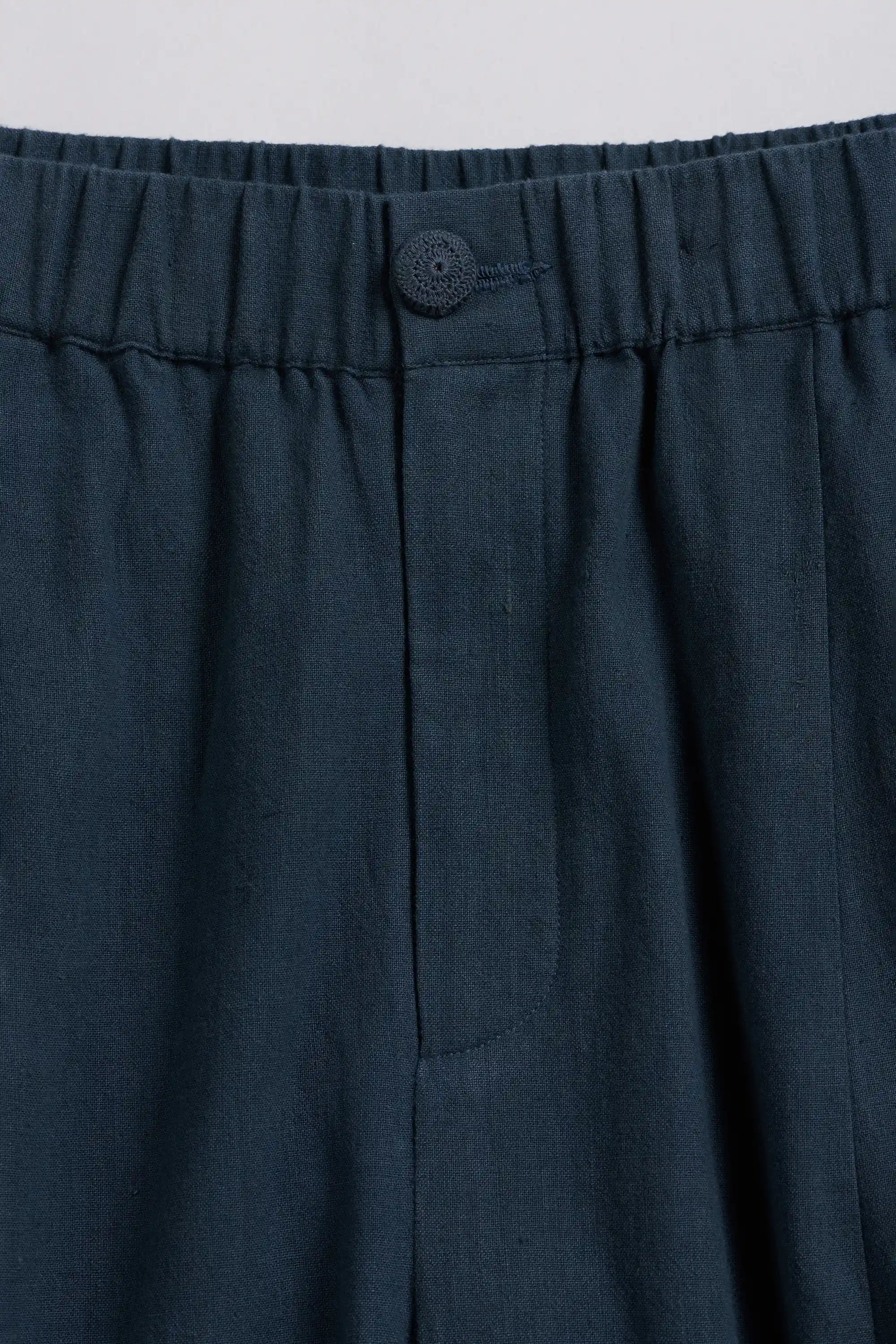 Close-up of black indigo pleated trousers showing the elastic waistband, button closure, and fabric texture.