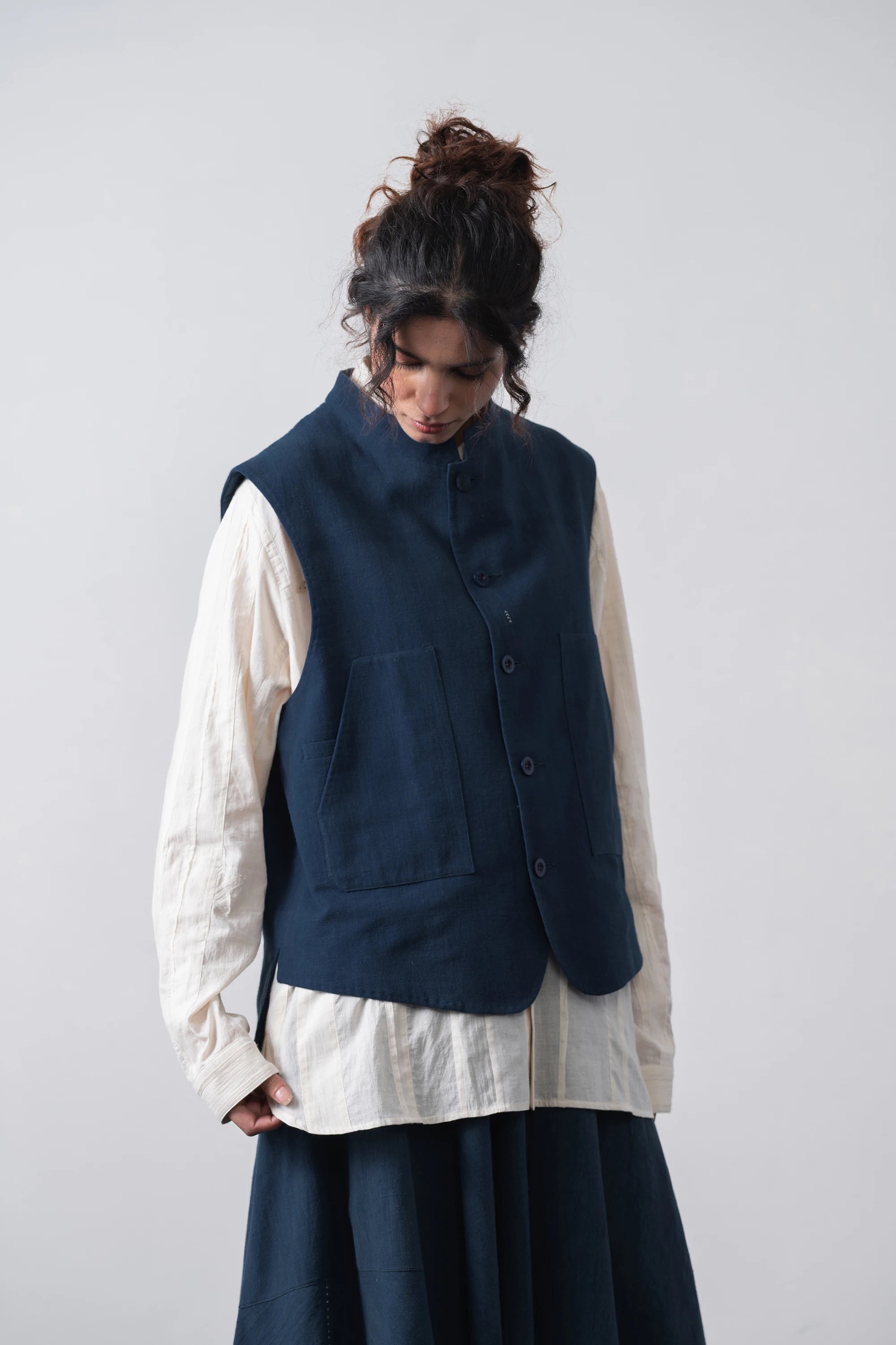 A model wearing a space blue vest over a cream-colored linen shirt and a skirt, standing against a light grey studio background.