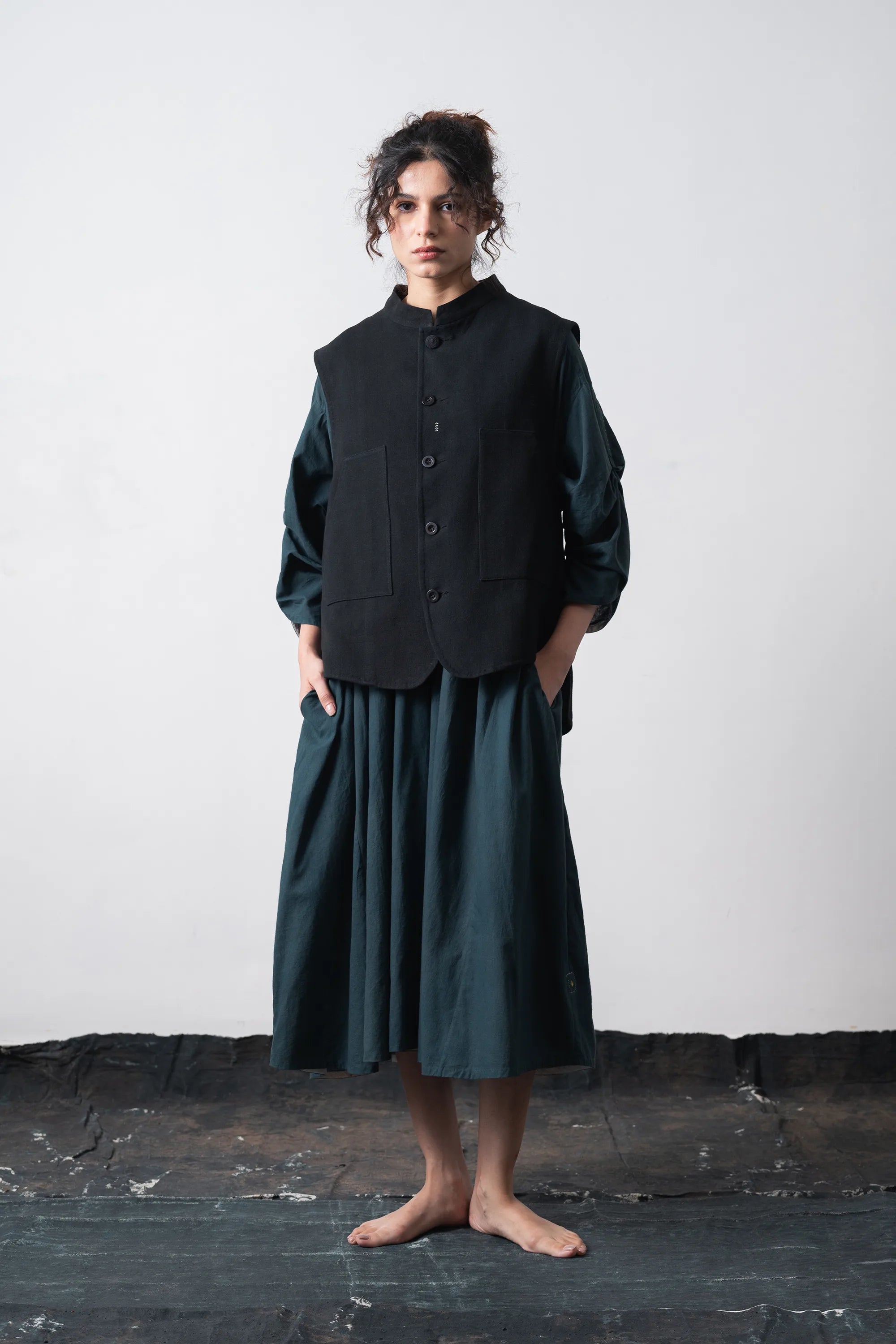 A model stands barefoot against a light grey wall and dark floor, wearing a black vest over a long-sleeve top and matching pleated midi skirt.