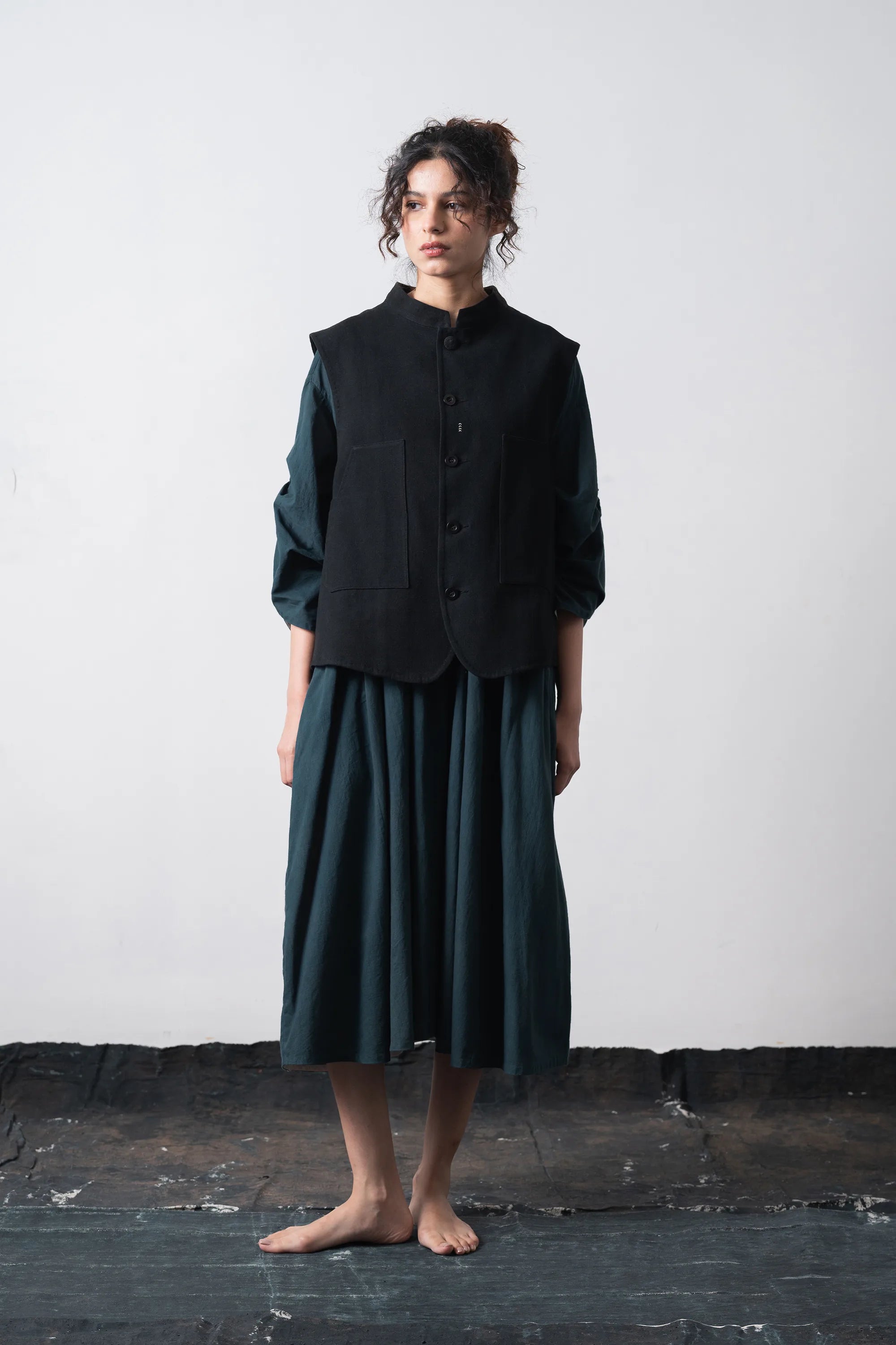 A model stands barefoot against a plain light grey wall, wearing a black vest over a long-sleeve shirt and a matching pleated midi skirt.