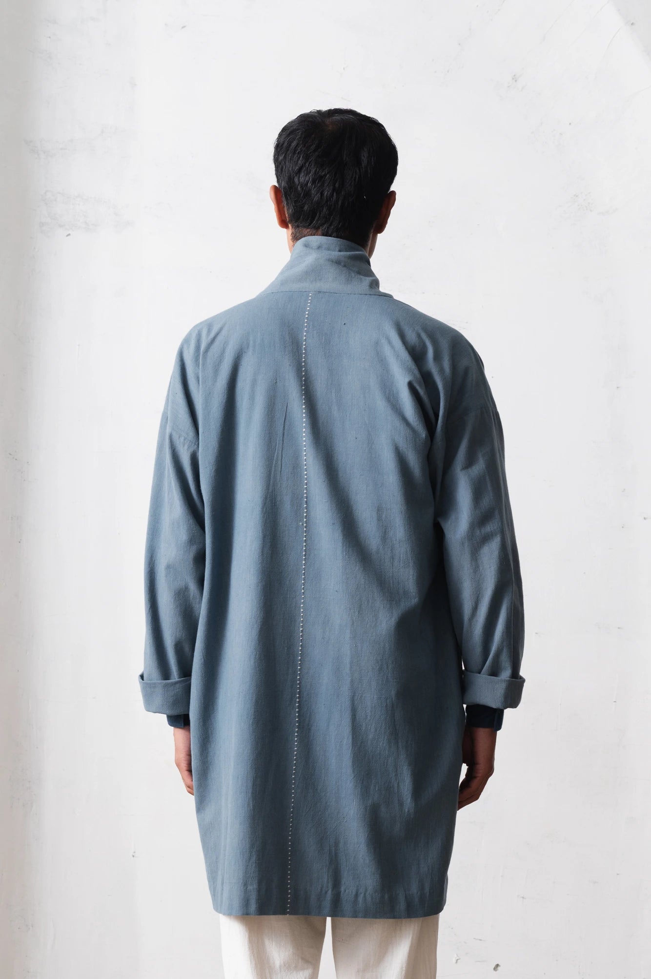 Back shot of a man wearing a long, light blue shacket with rolled-up sleeves against a light grey background
