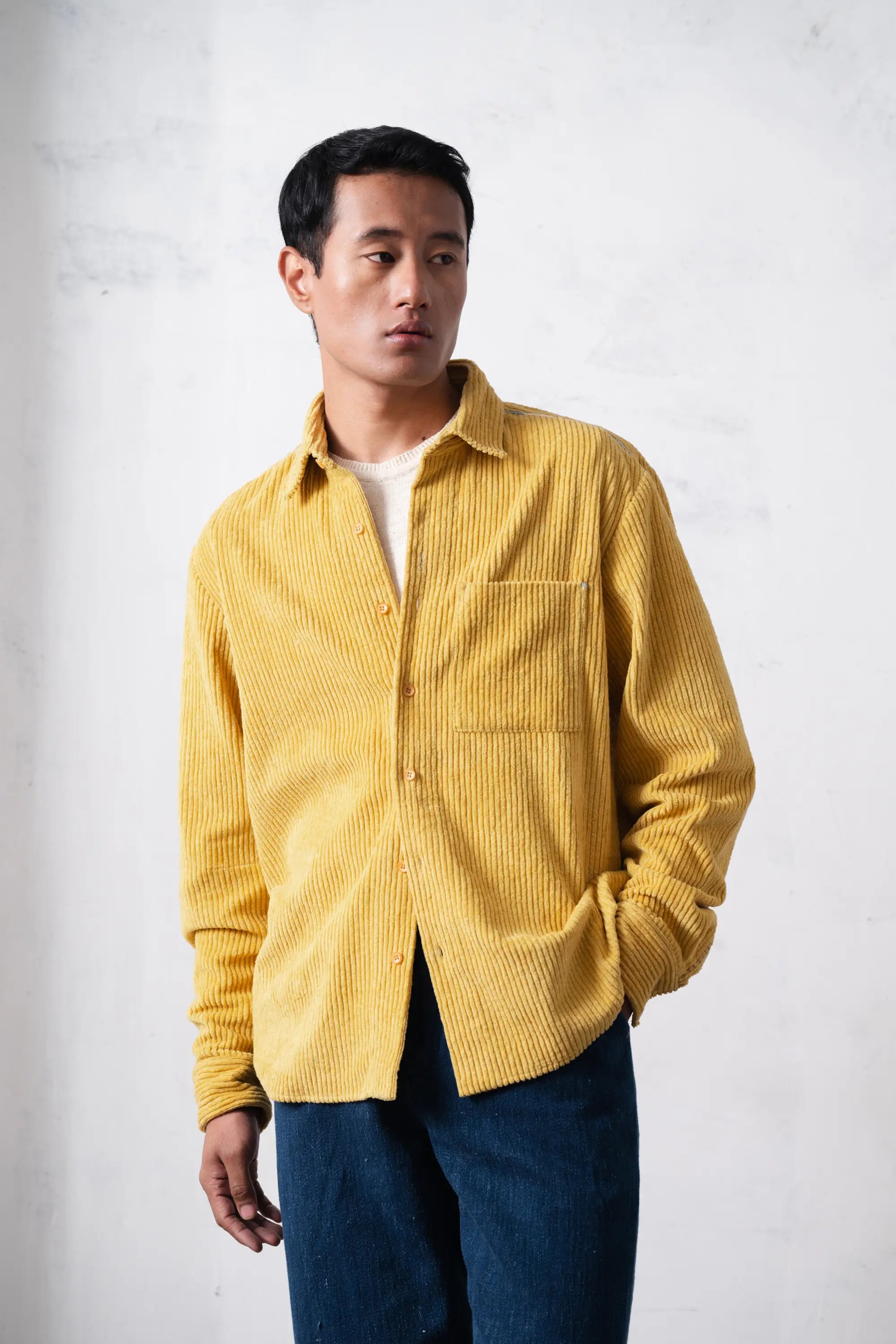Man wearing a yellow corduroy shirt over a white t-shirt with blue pants against a light grey background