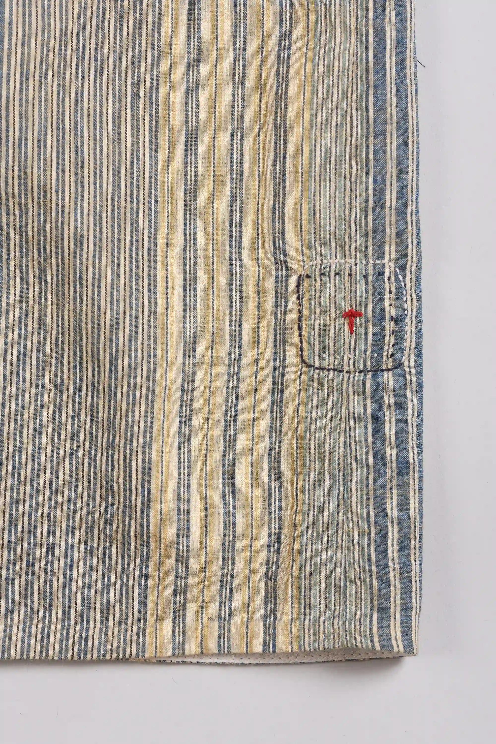 Close-up of a striped textile, with vertical blue, beige, and grey stripes and a small red embroidered detail on a white background.