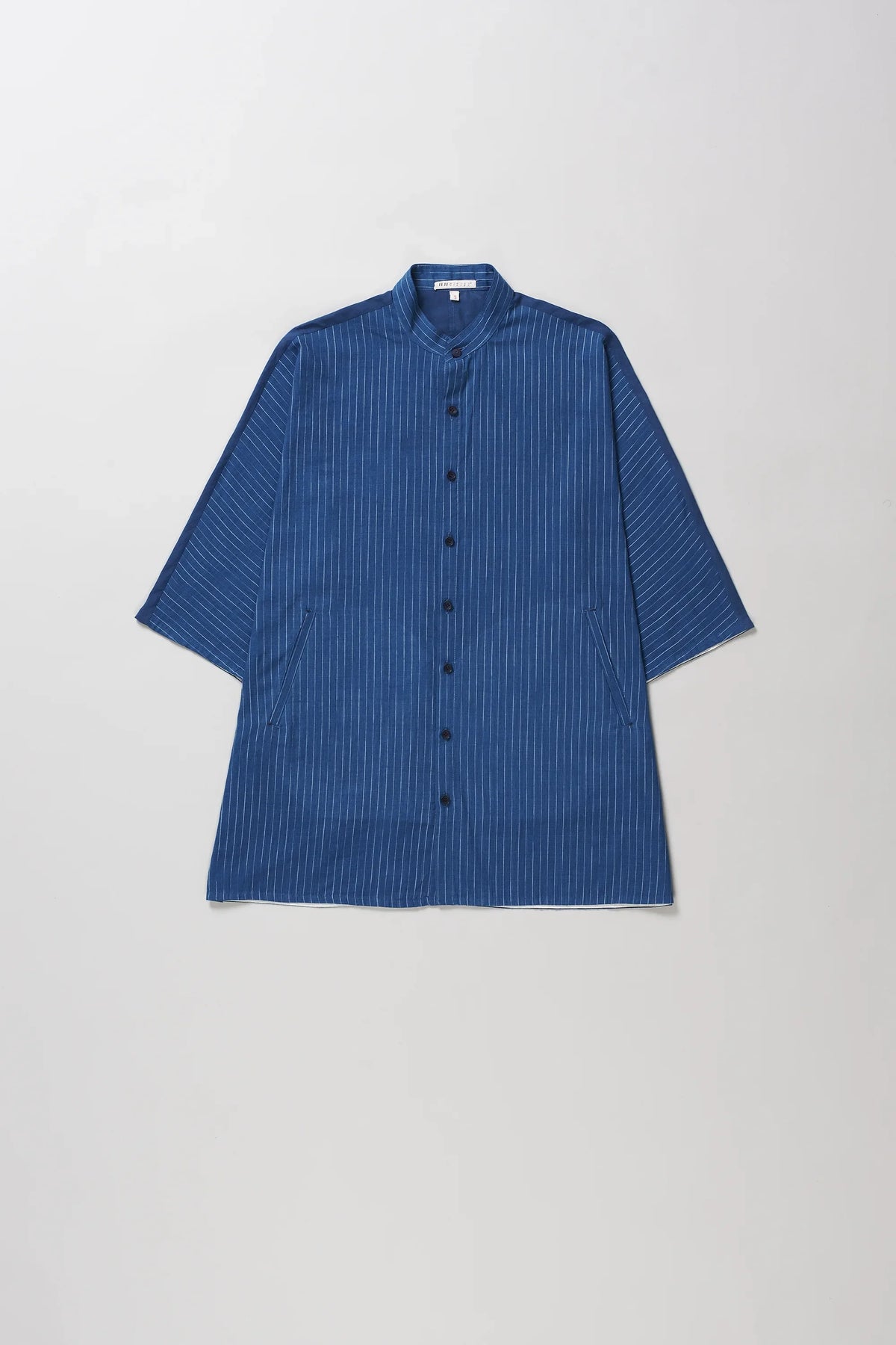 A indigo short-sleeve button-up shirt with vertical white pinstripes, a mandarin collar, and black buttons, laid flat against a light grey background.