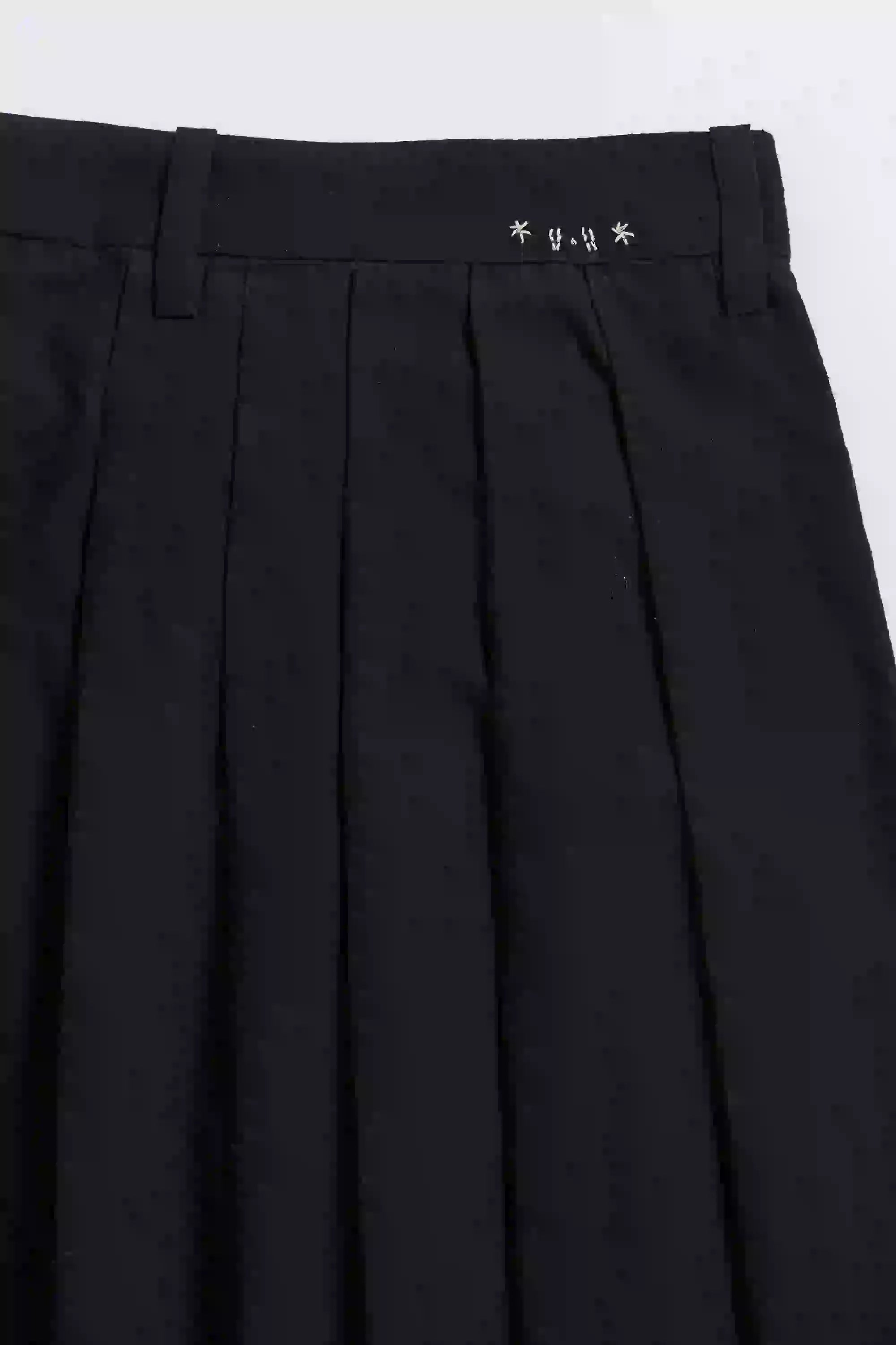 Black pleated pants with a brand logo on the waistband against a grey background