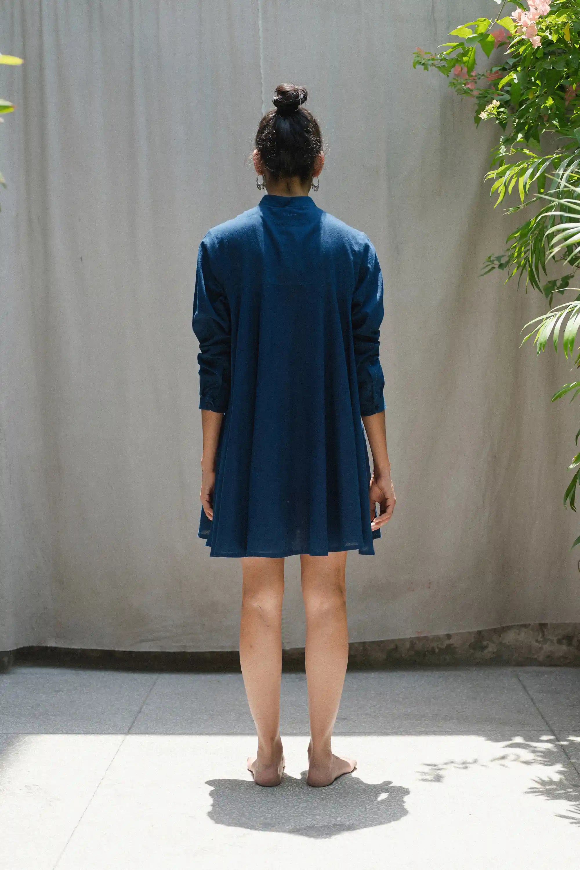 Back shot of a parson wearing a blue dress standing against a plain wall with plants on either side.