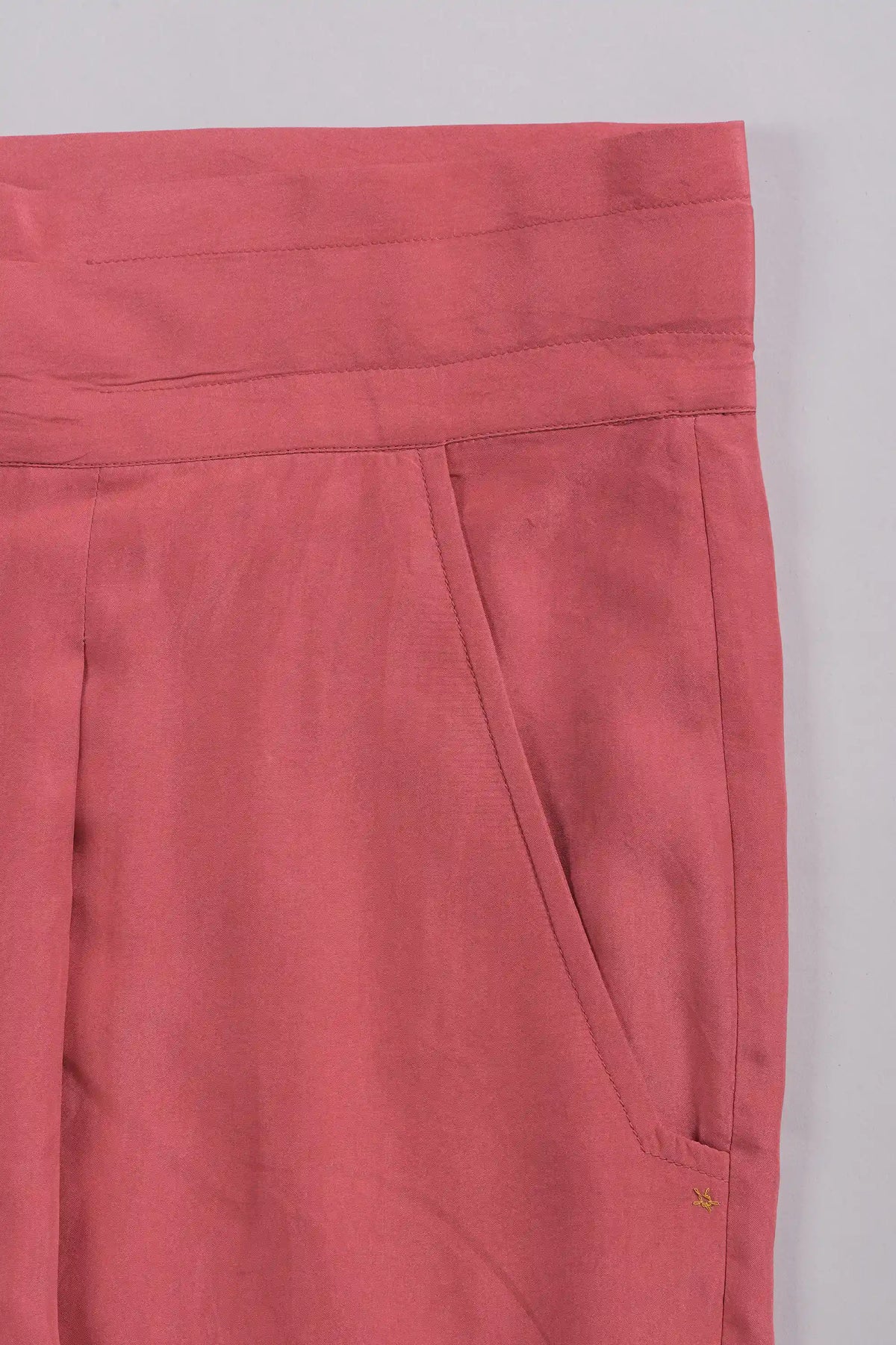 Close-up of a dusty rose pink garment, showing the waistband, front seam, and side pocket detail against a light grey background.