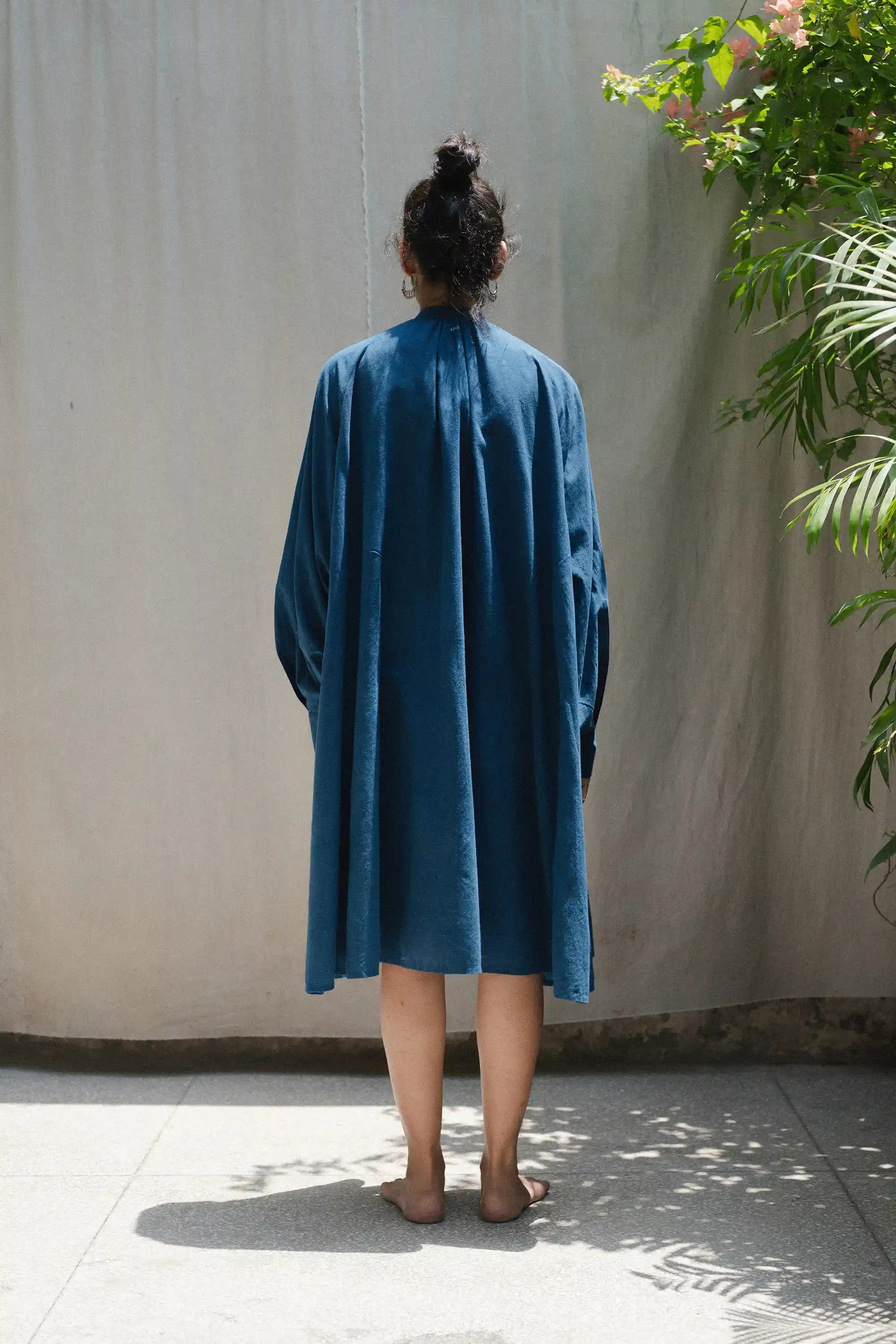 Person wearing a blue dress standing against a plain wall with plants around.