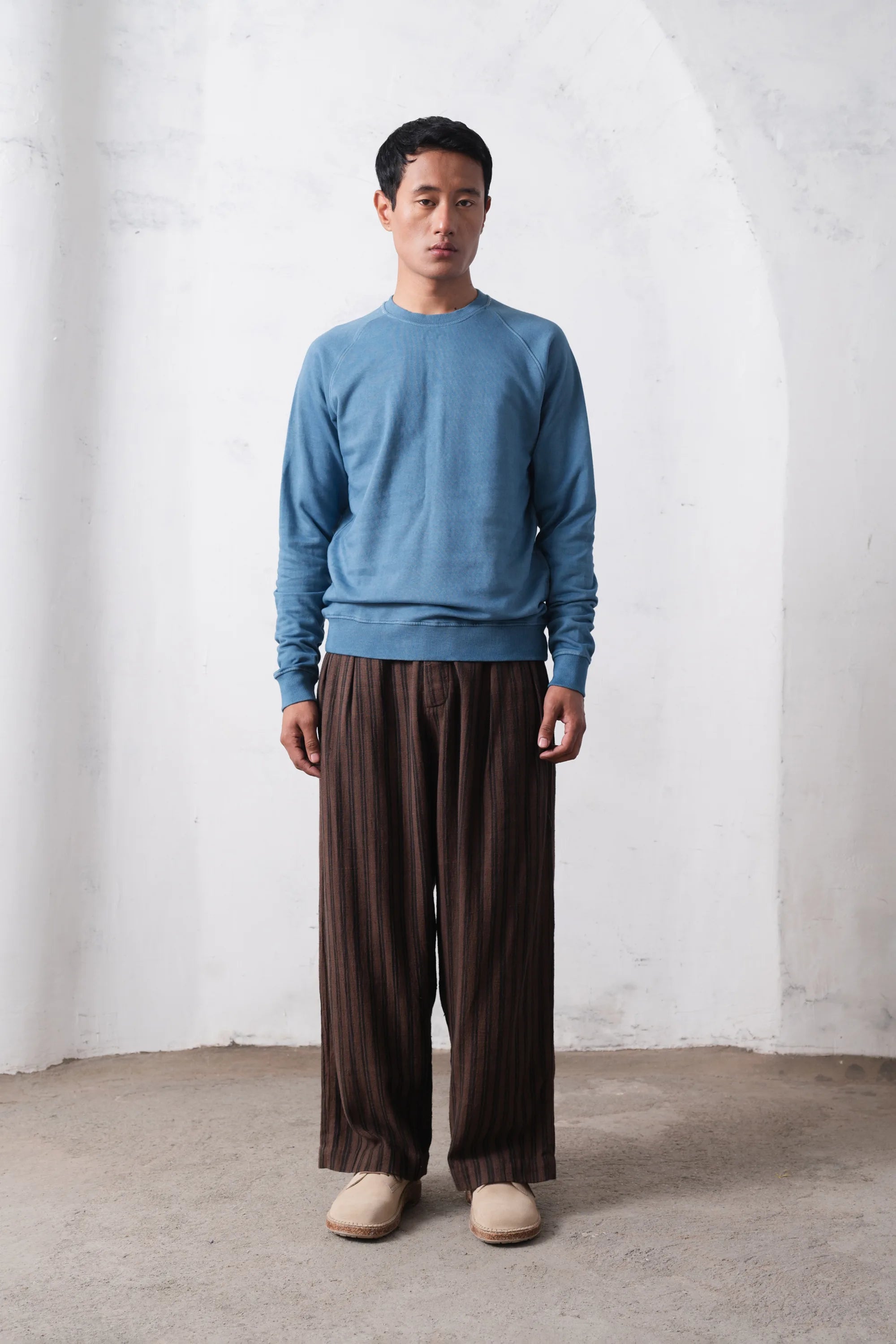 Man wearing a blue sweater and brown pants standing against a light grey wall