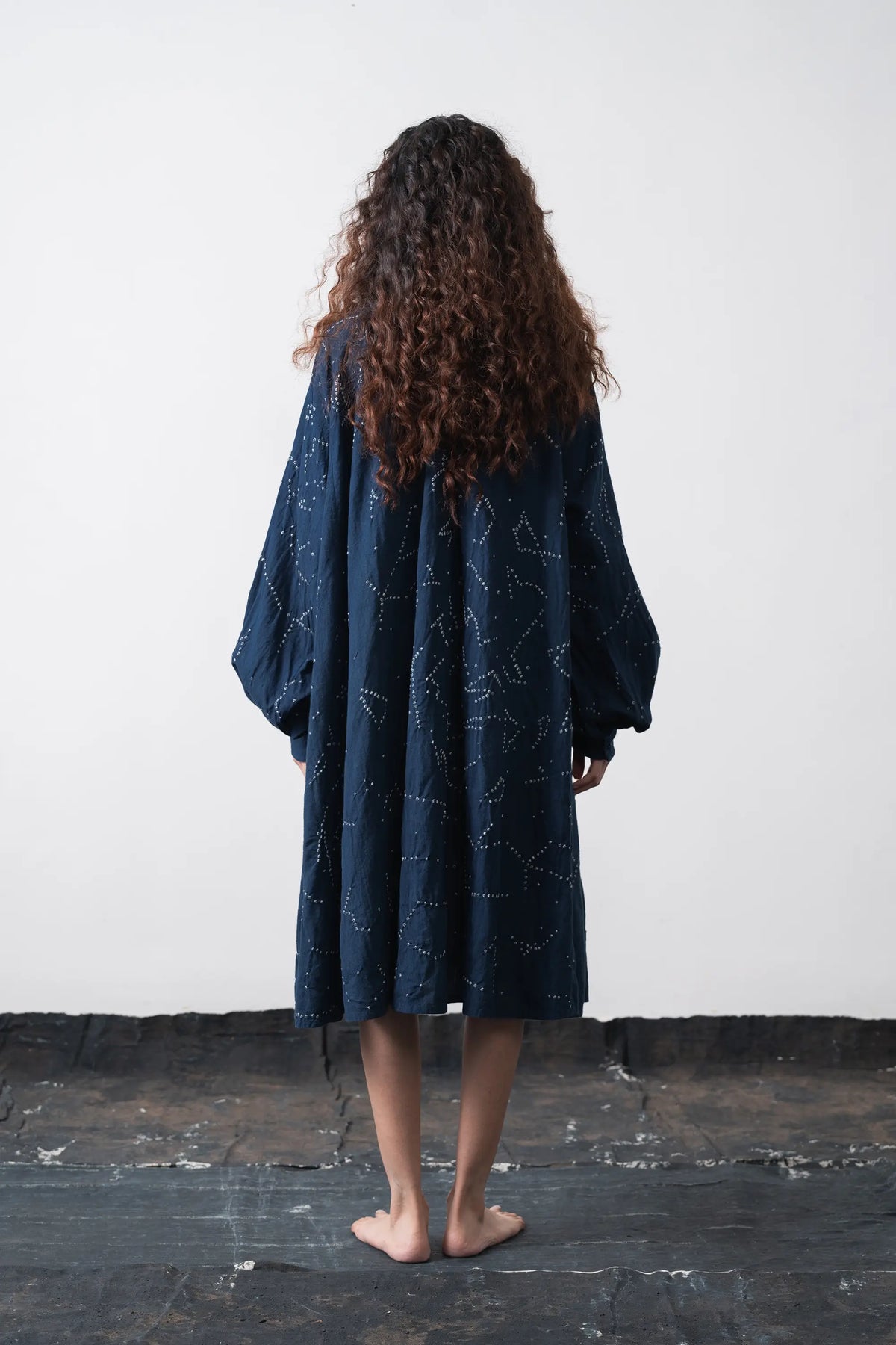 A woman stands barefoot from the back, wearing a loose, space blue dress standing against the wall.