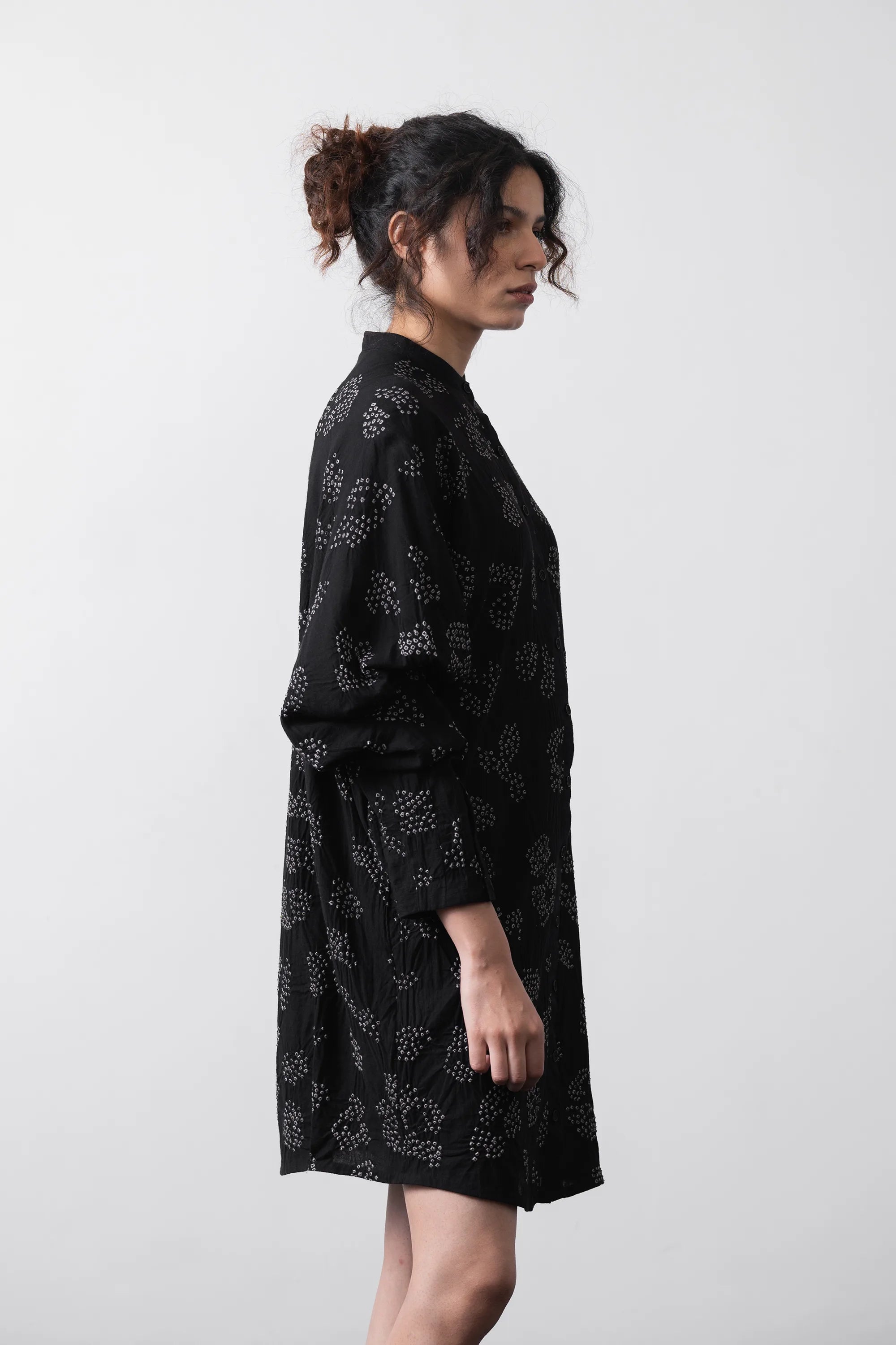 A woman wearing a black long-sleeve tunic dress with a white floral pattern, shown from a side profile against a plain grey background.