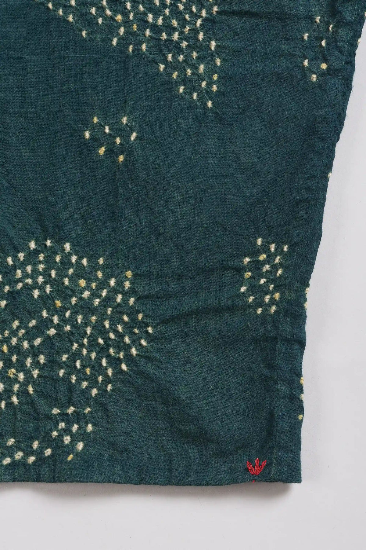 A close-up of a bottle-green fabric with white tie-dye bandhani dot, finished with a small red embroidered logo on the bottom hem.