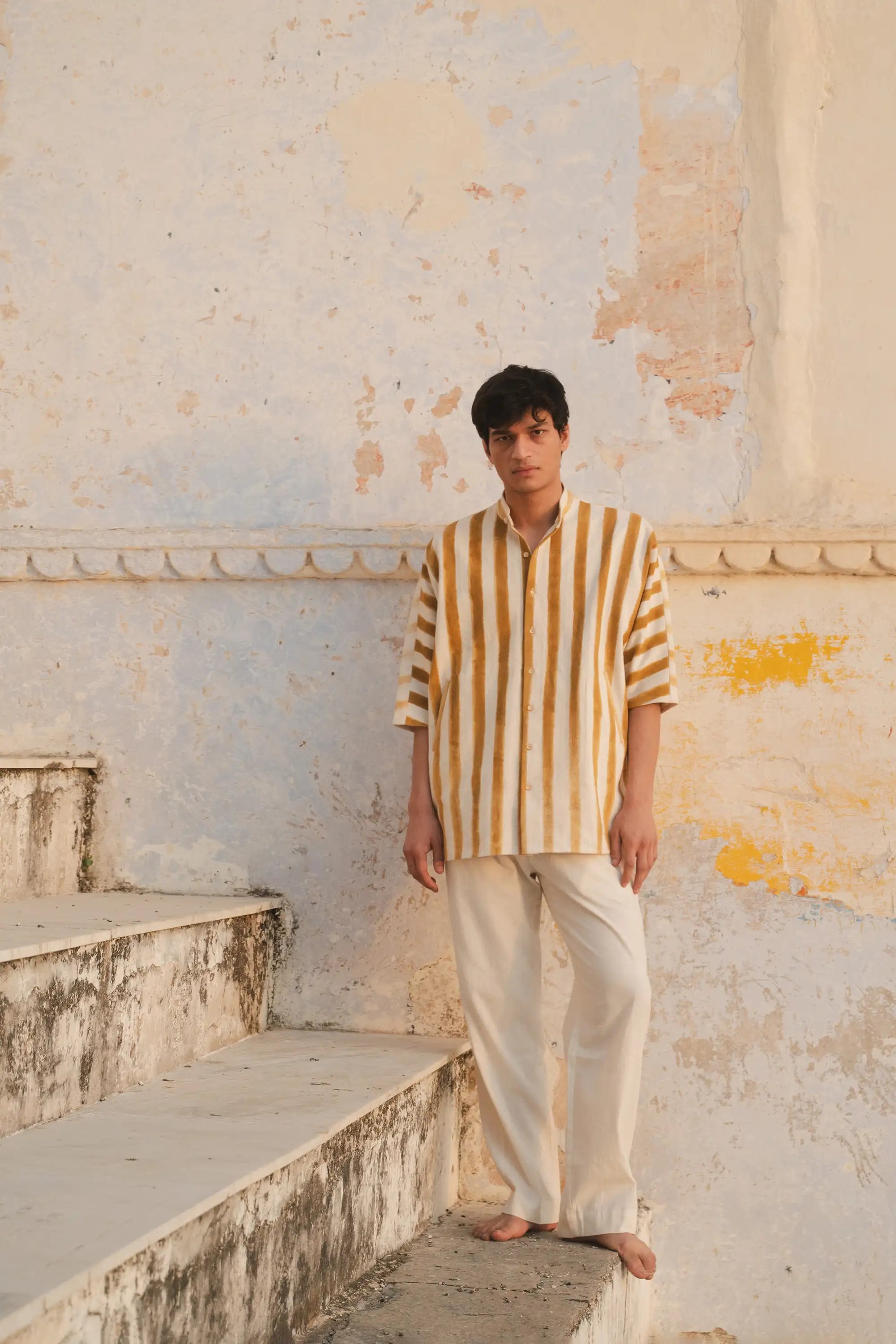 Person wearing a striped shirt and light pants against a textured wall.