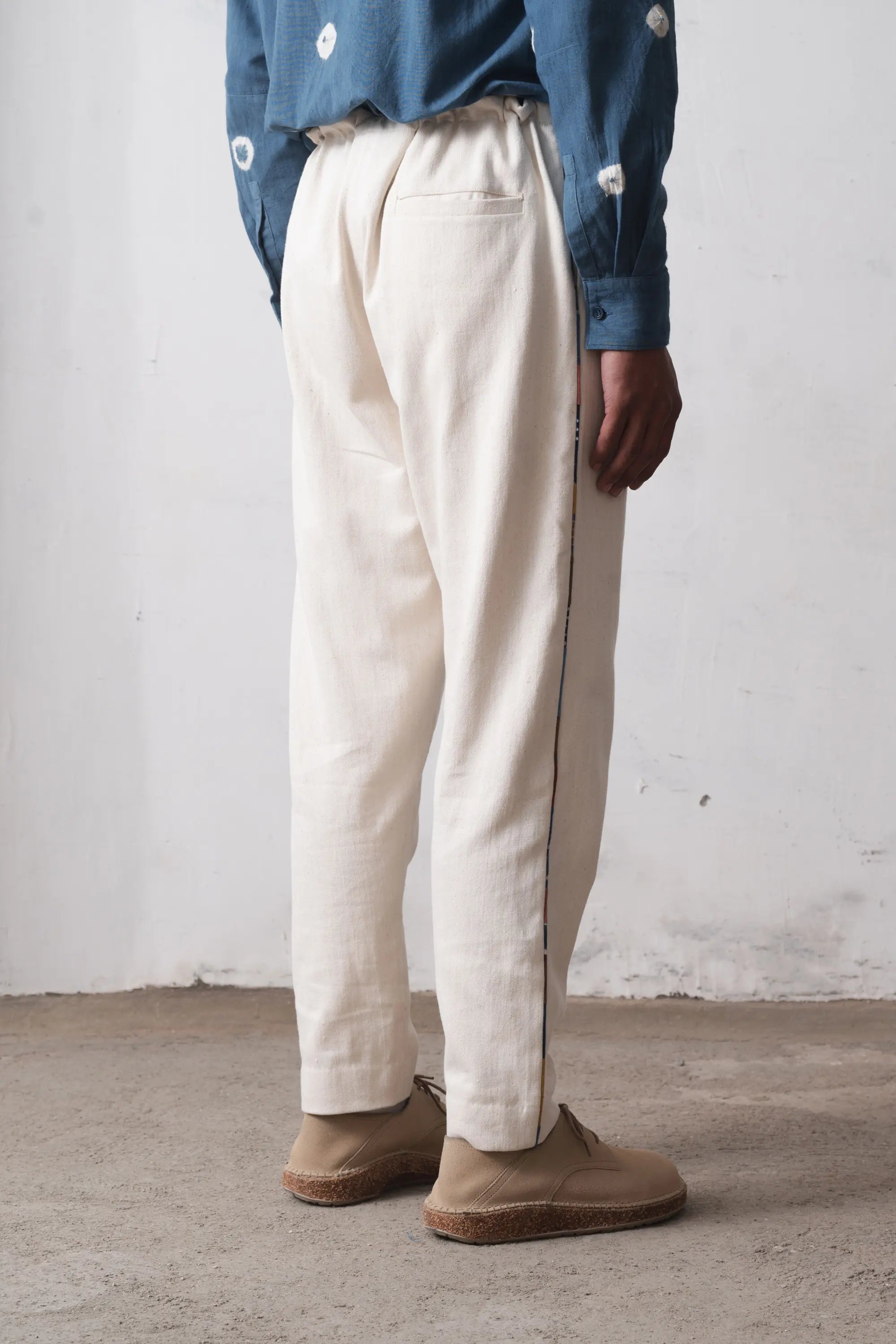 A back shot of a person wearing ecru pants and a blue shirt against a white wall