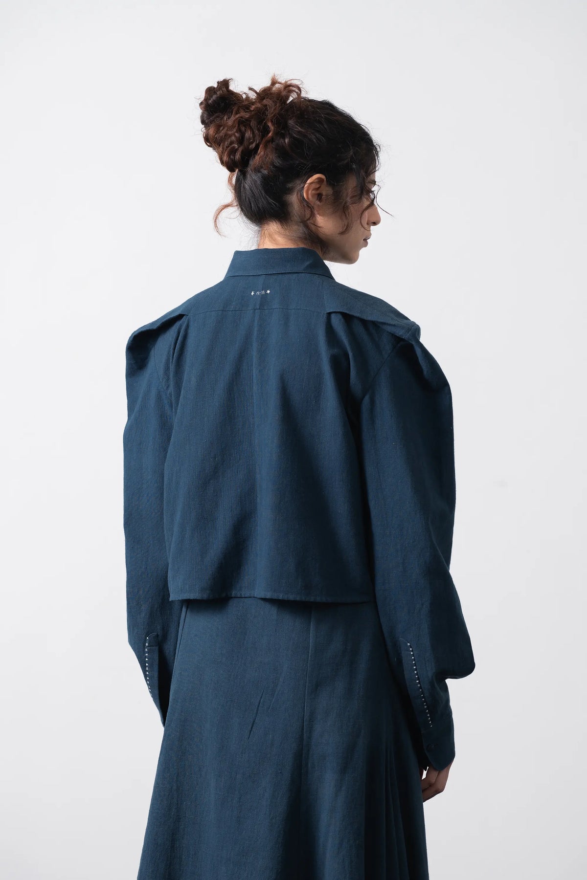 A model shown from the back wearing a space blue long-sleeve shirt with dramatic shoulder puffs and a matching skirt, set against a plain light grey background.