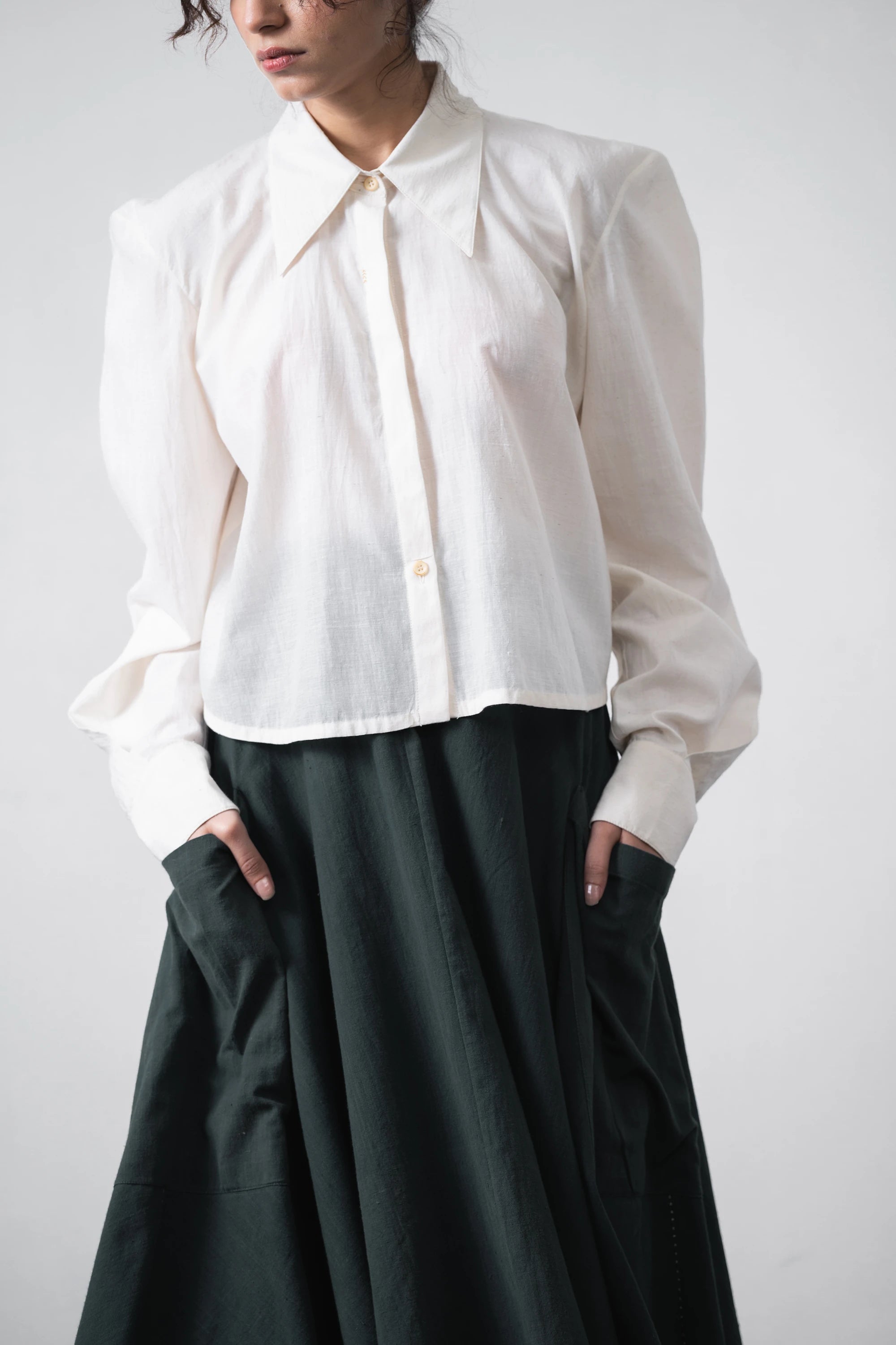 A model wearing a white long-sleeve button-up shirt with a pointed collar and a dark green skirt, shown from the neck down against a light grey background.