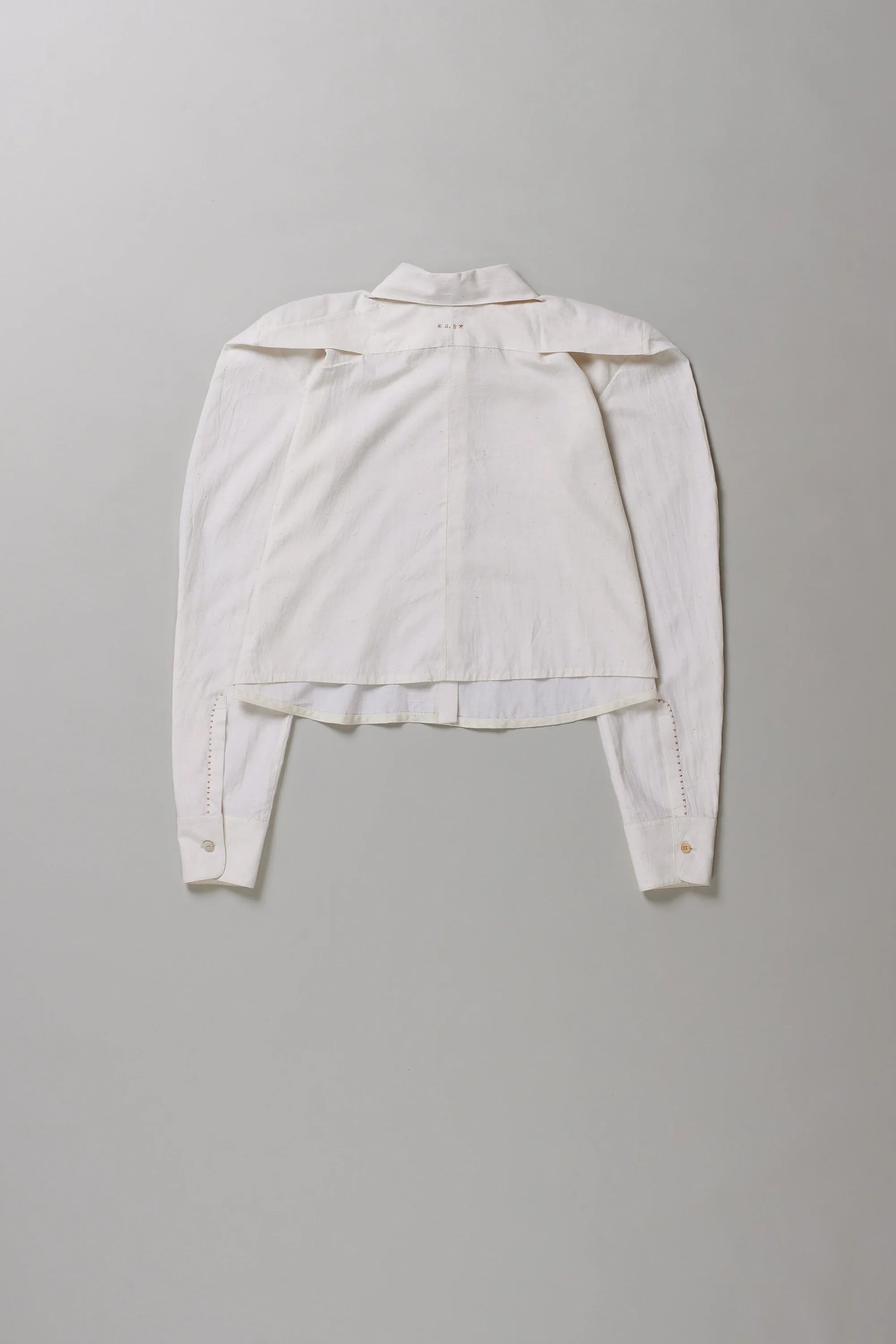 Back shot of off-white long-sleeve cropped shirt with structured shoulders and a high-low hem, laid flat against a neutral gray background.