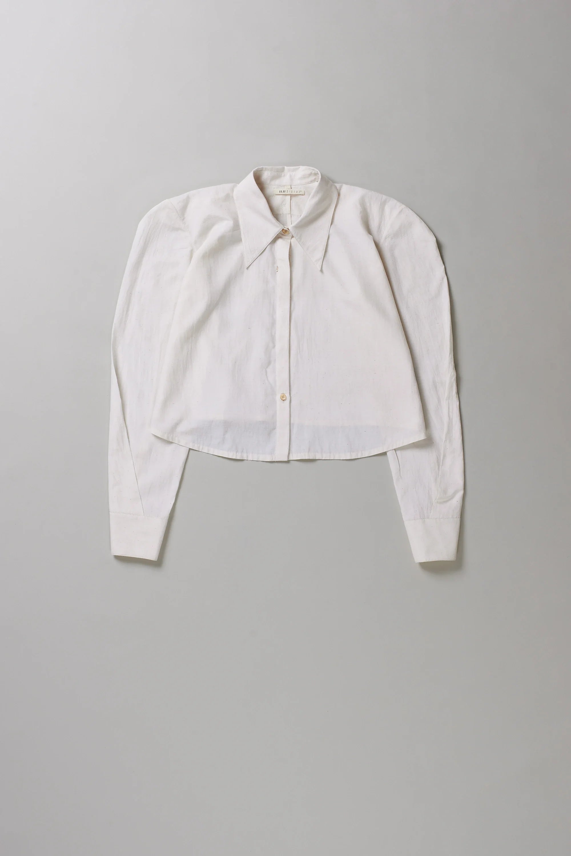 A cropped long-sleeve button-up shirt in off-white, laid flat against a neutral light gray background.