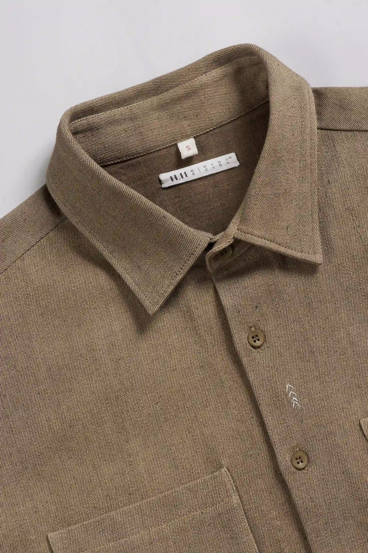 A close-up of a brown textured linen shirt with a pointed collar, button-down front, and a visible brand label inside the collar.