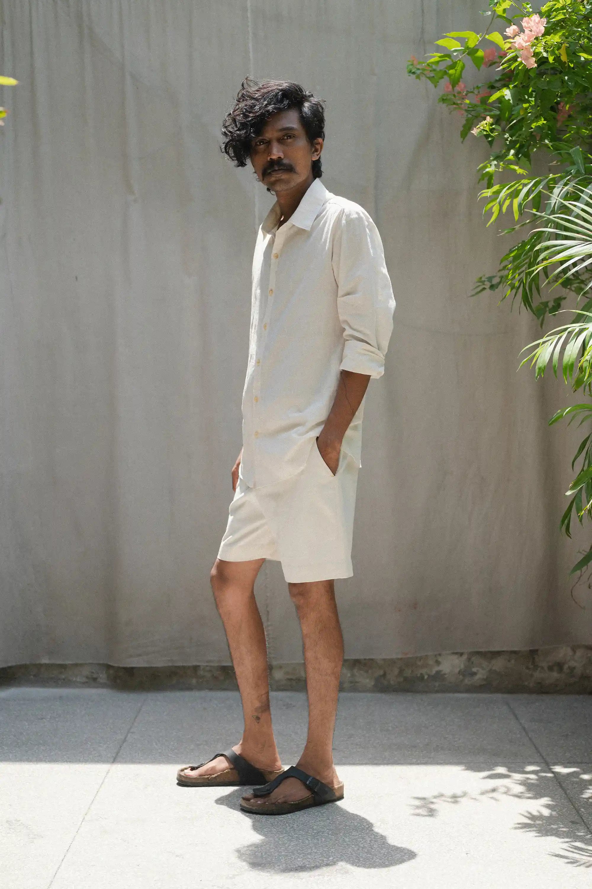 Man in a ecru shirt and shorts standing against a plain wall with plants around.