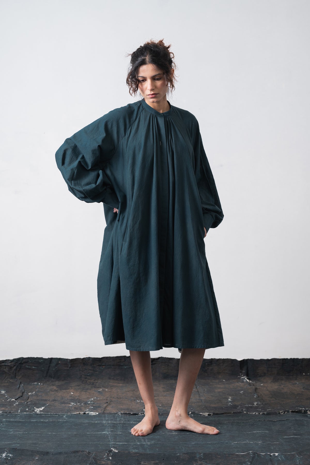 Woman stands barefoot against a wall wearing a loose, oversized black indigo dress with voluminous long sleeves and a gathered neckline.
