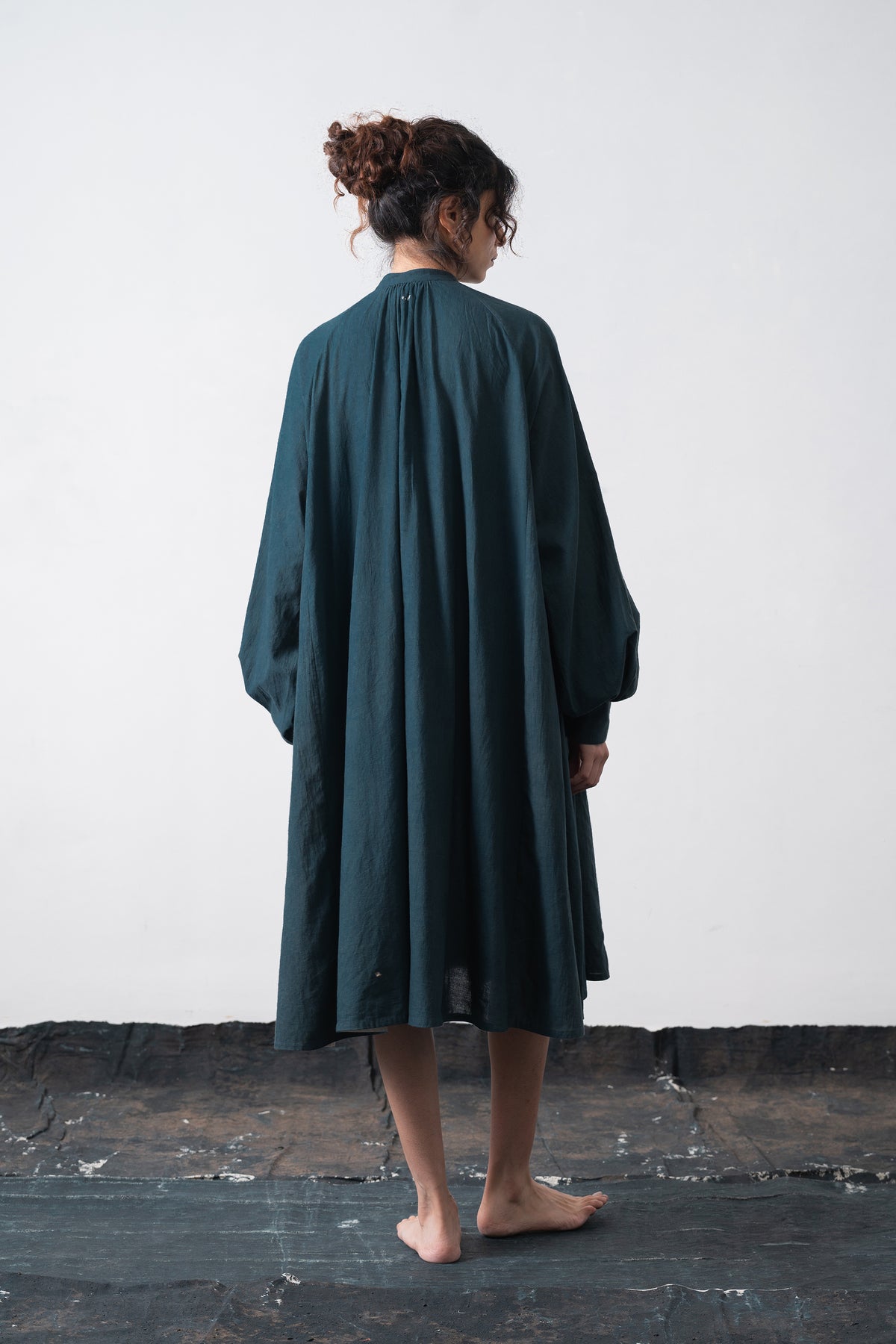 A woman viewed from behind wearing a black indigo long-sleeved dress, standing barefoot on a dark wooden floor against a plain white wall.