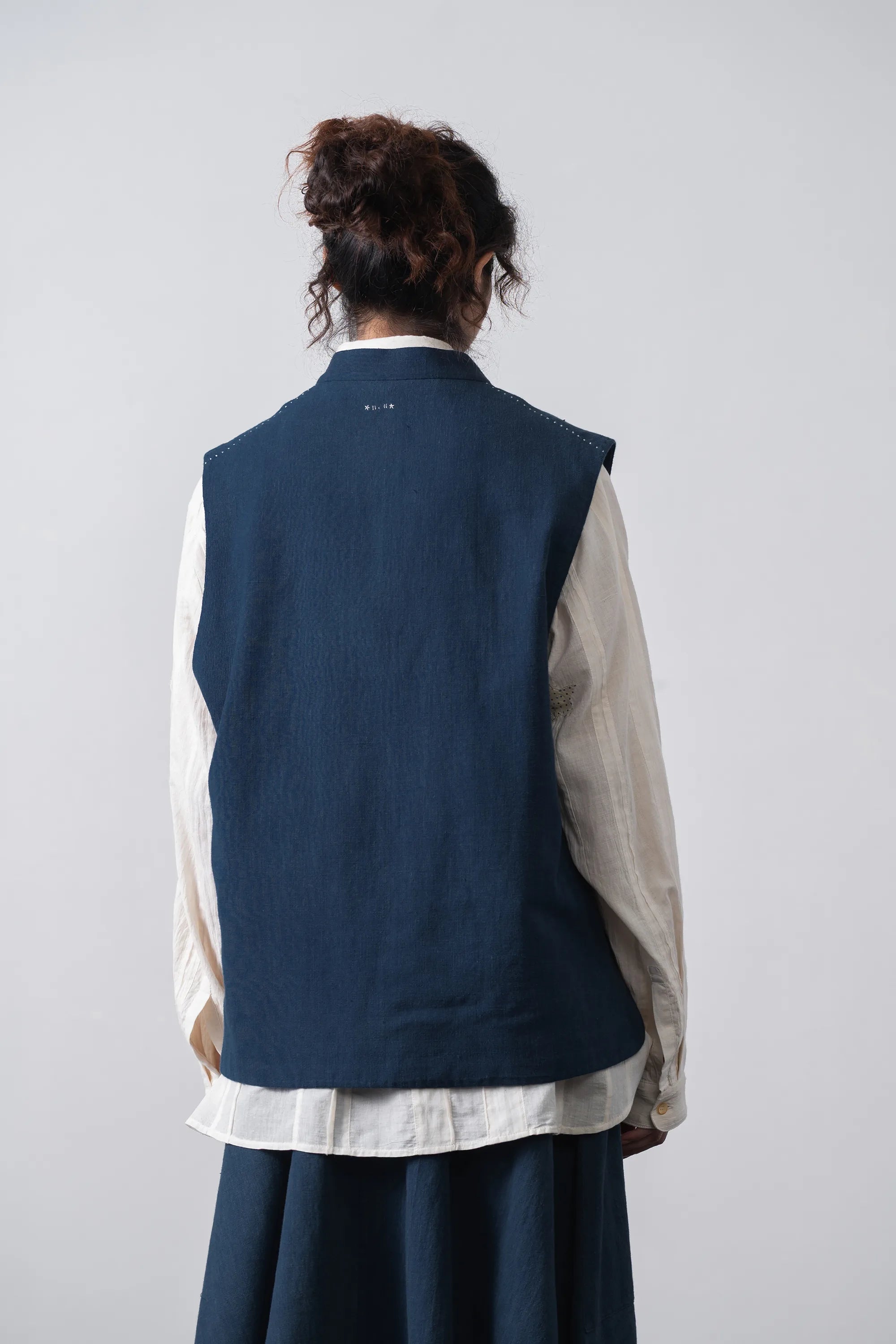 Woman shown from the back wearing a sleeveless navy vest over a shirt and skirt, photographed against a plain light grey studio background.