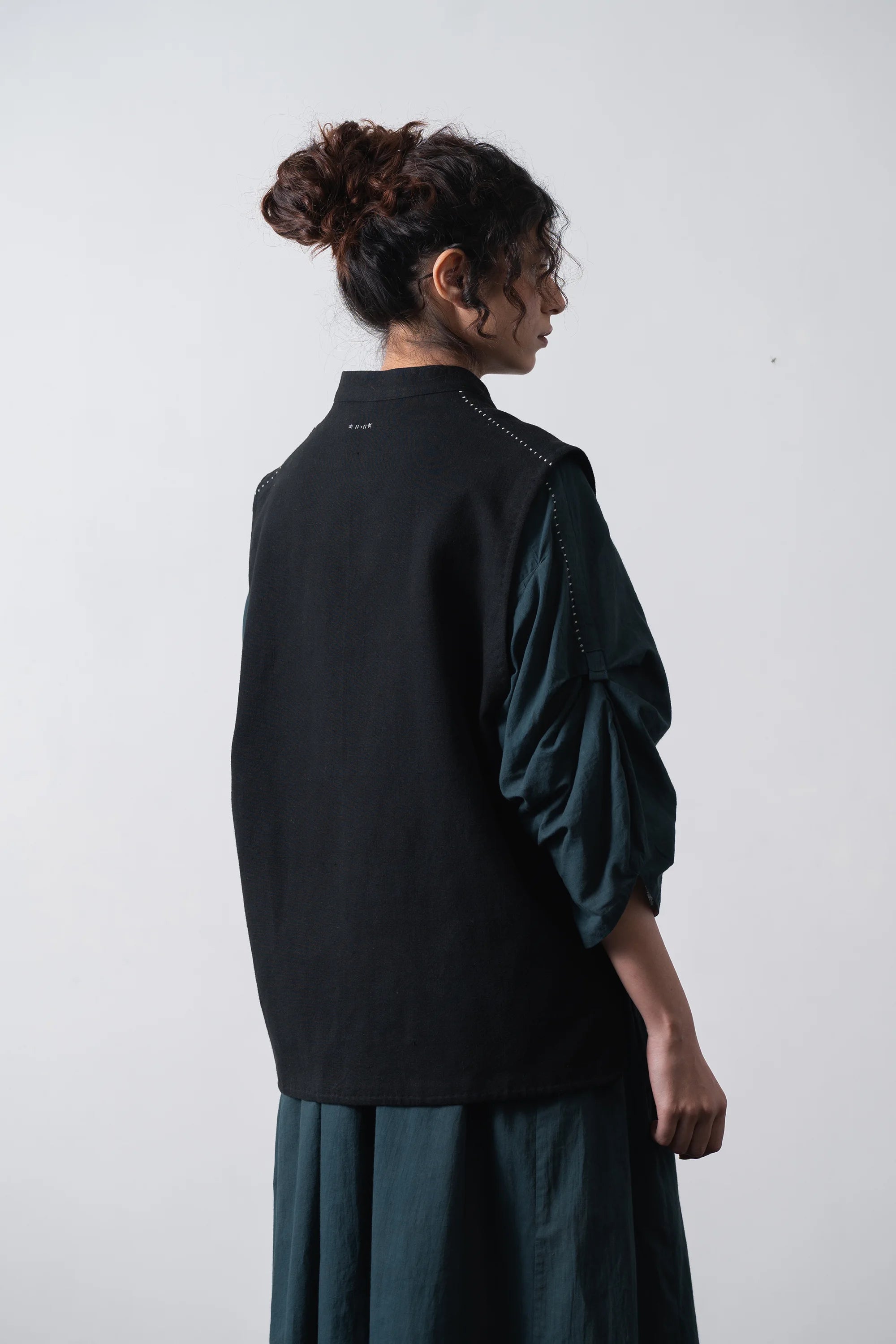 A woman shown from the back wearing a black sleeveless vest over a shirt and matching skirt, photographed against a plain light grey studio background.