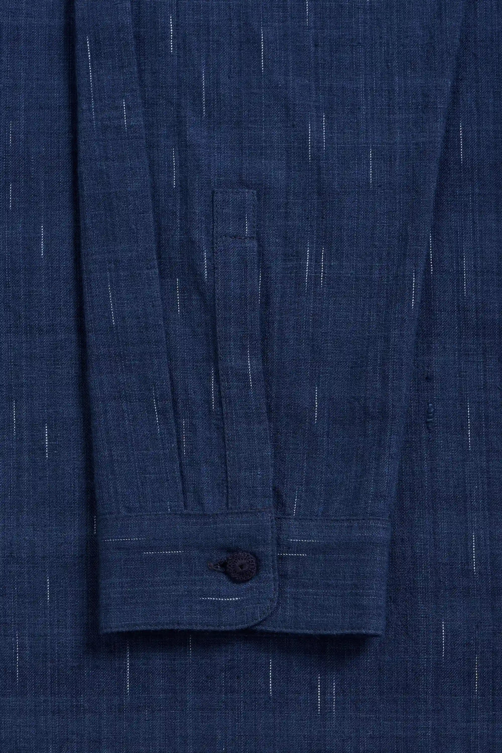 Close-up of a medium indigo textured fabric with fine vertical white stripe details and a buttoned cuff