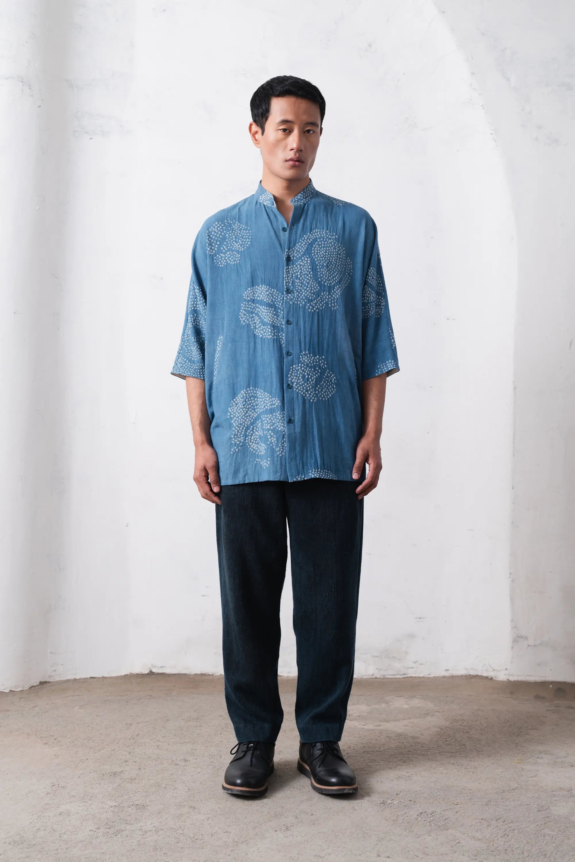 Man wearing a blue bandhani shirt and dark pants against a light grey wall.