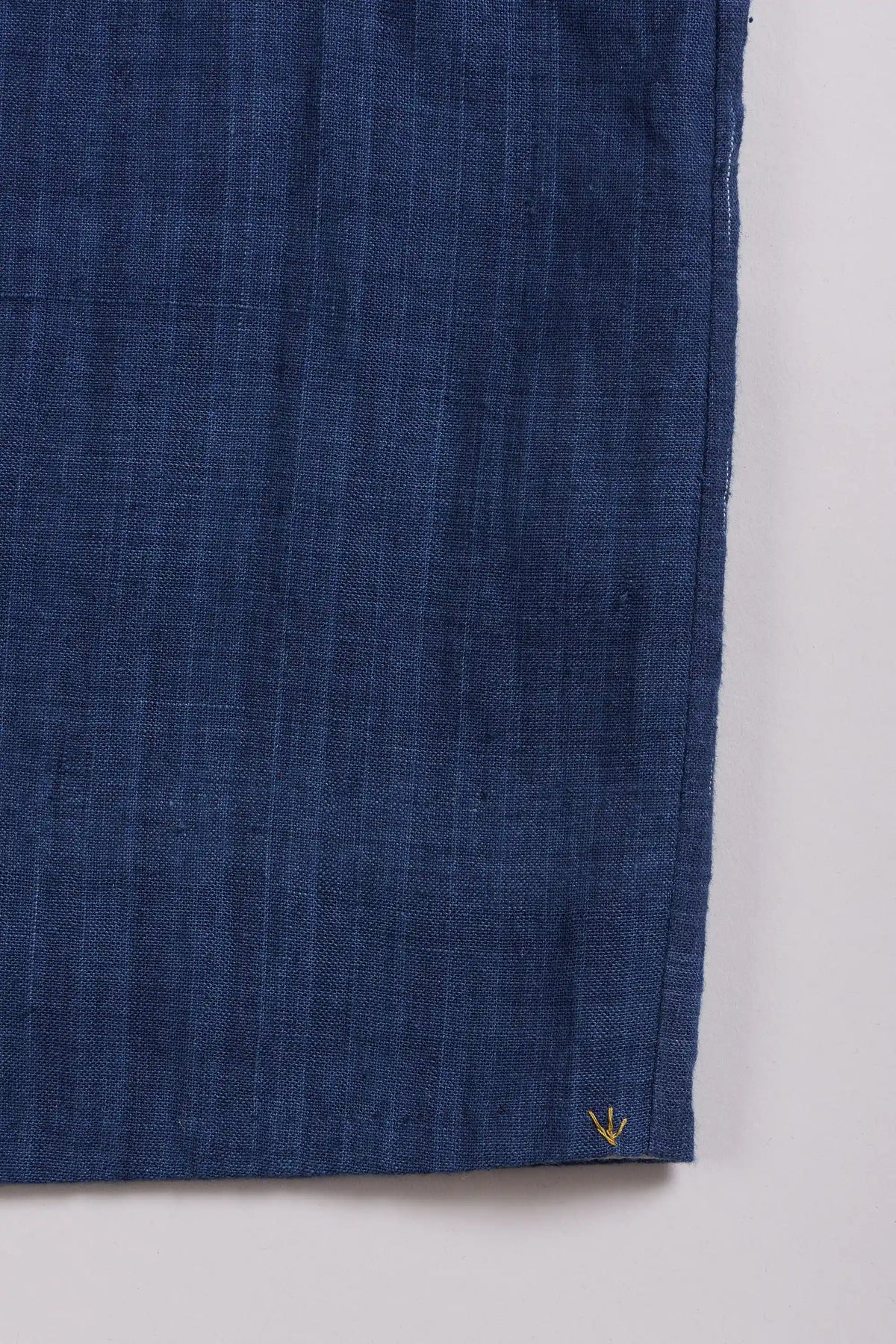 A close-up of a indigo textured fabric, likely cotton, showing a fine woven grid pattern and a small yellow arrow stitched near the bottom right corner, set against a plain light grey background.
