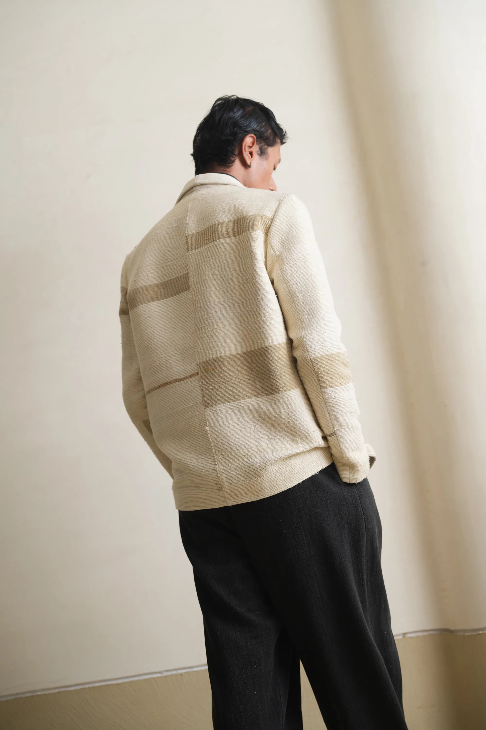 Back shot of a man wearing a beige cardigan over a black shirt and pants against a light grey wall