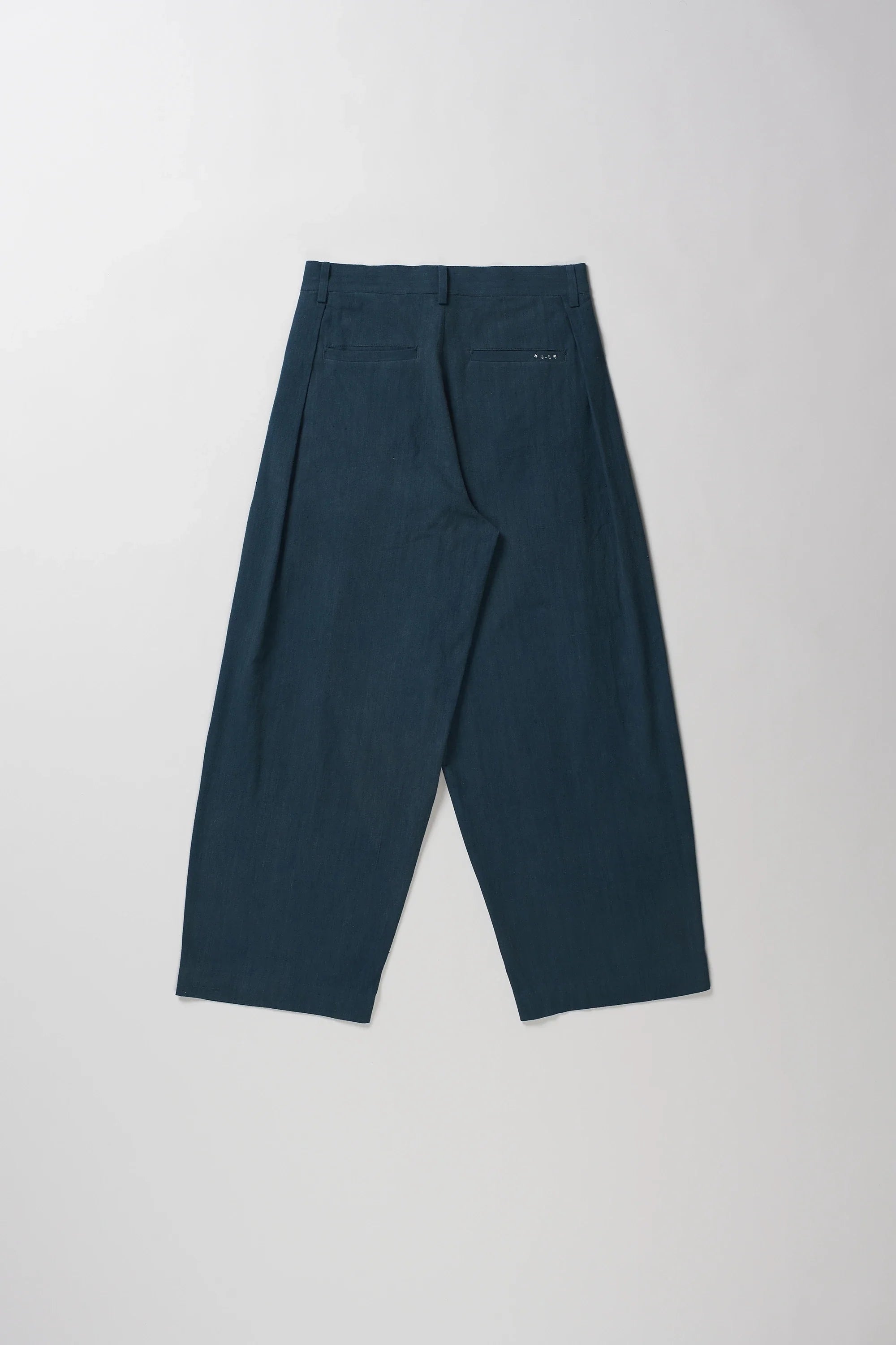 Back shot of pair of black indigo pleated trousers laid flat against a light gray background.