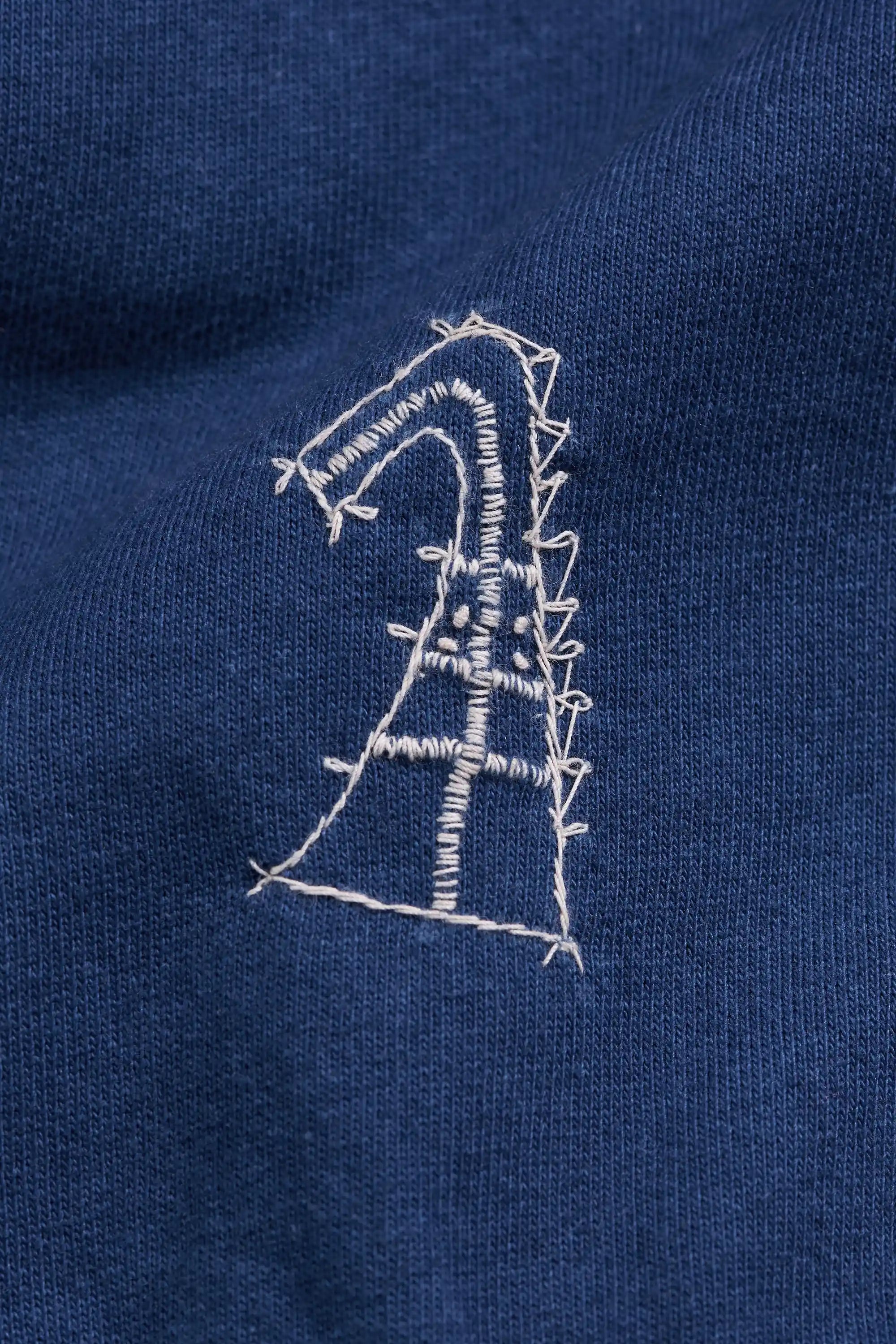 Close-up of a medium indigo knit fabric with a small white embroidered graphic resembling a stylized letter or abstract shape.