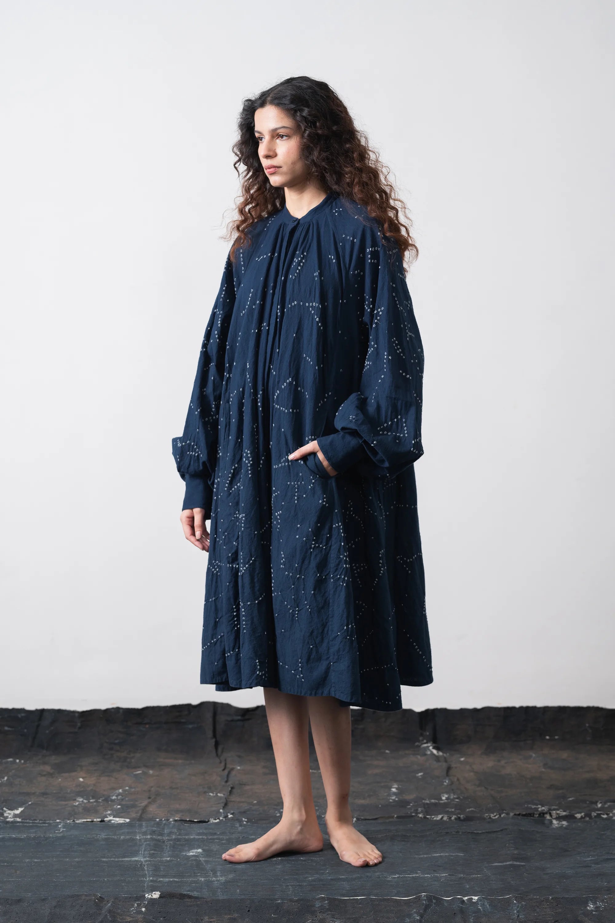 A woman stands barefoot against a grey wall, wearing a loose, space blue long-sleeve dress with a subtle celestial star pattern.