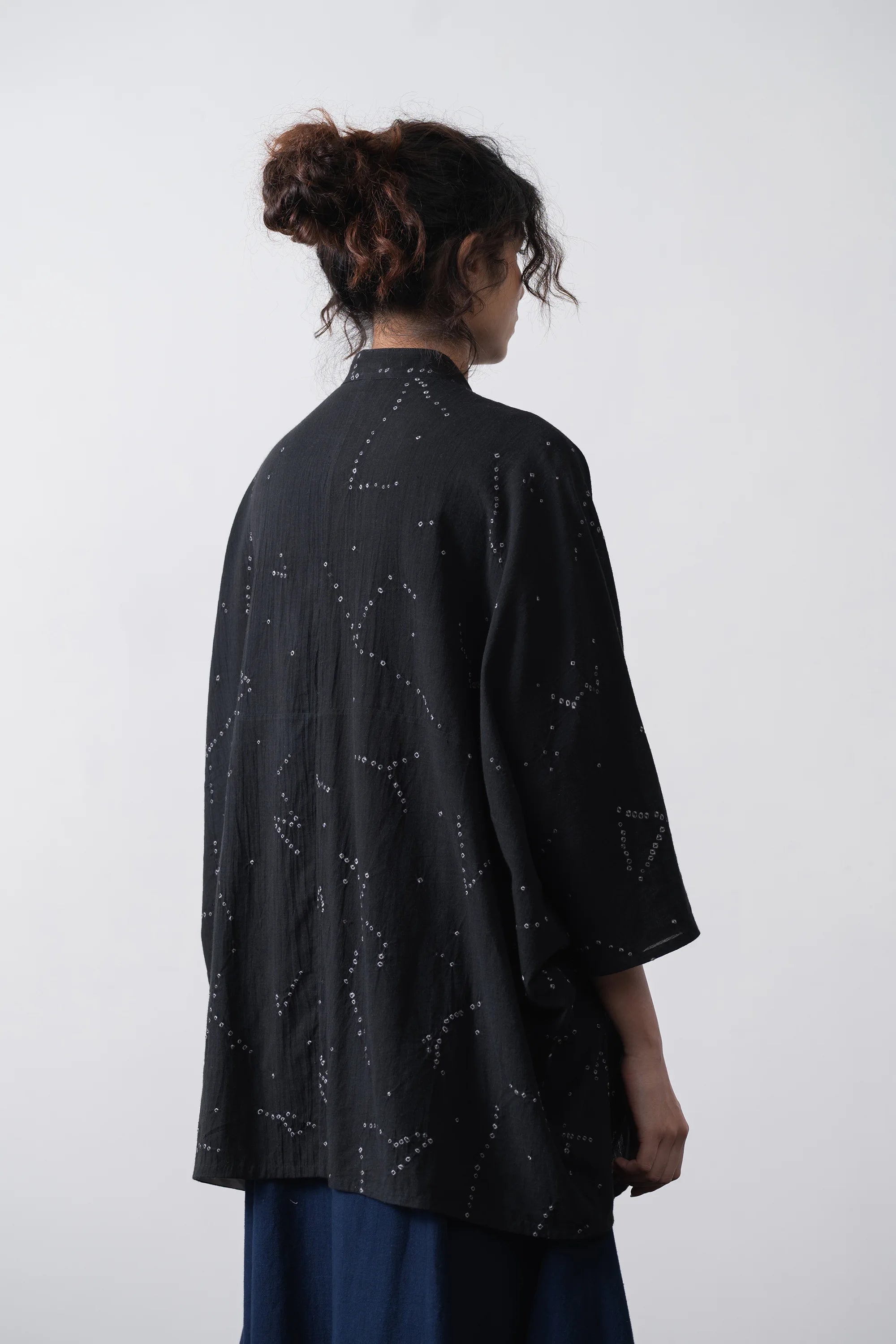 A woman shown from the back wearing a loose black kimono-style shirt with shibori, paired pants, against a plain light grey background.