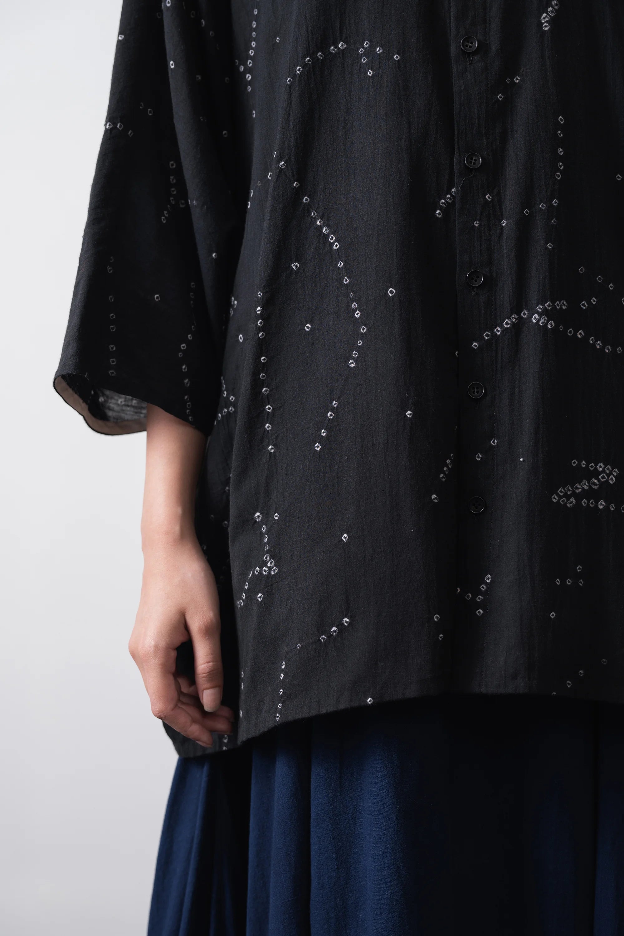 Close-up of a person wearing a black button-front shirt with shibori, paired skirt, against a plain grey background.