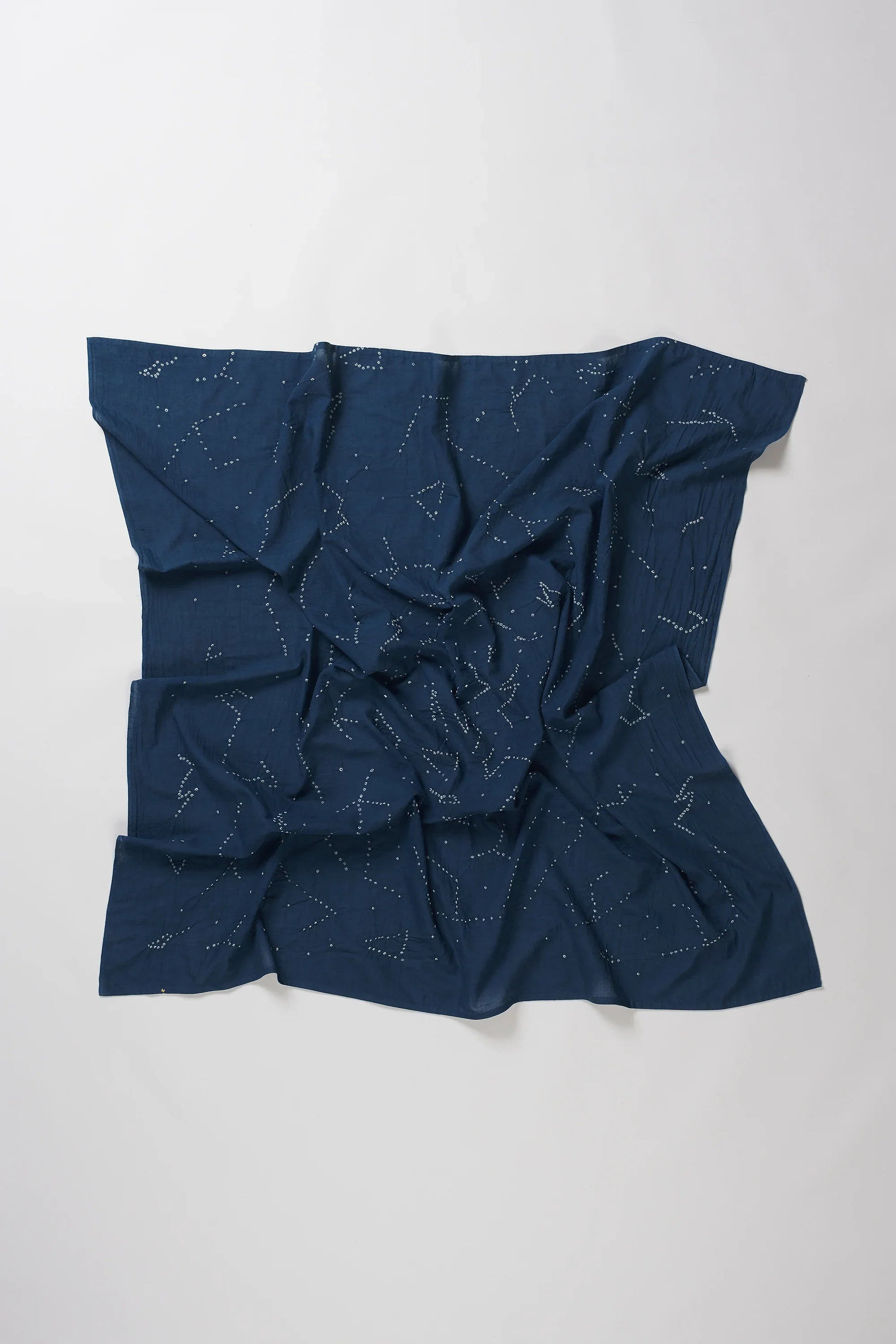 A space blue fabric accessory, likely a scarf or bandana, featuring a white celestial print of stars and constellations, laid out in a loose, crumpled square shape against a plain light grey background.