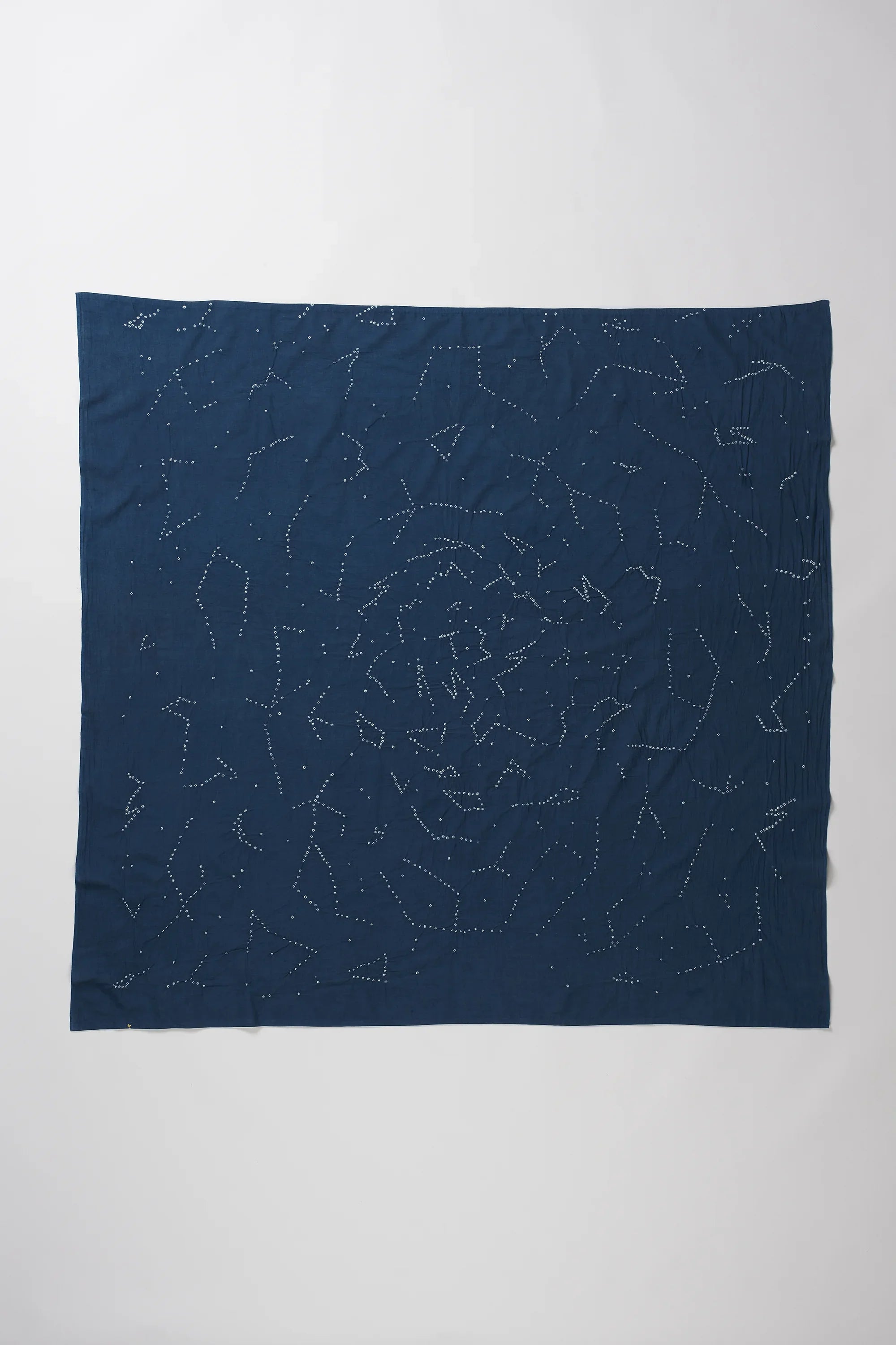 A space blue fabric accessory, likely a scarf or bandana, featuring a white celestial print of stars and constellations, laid out in a loose, crumpled square shape against a plain light grey background.