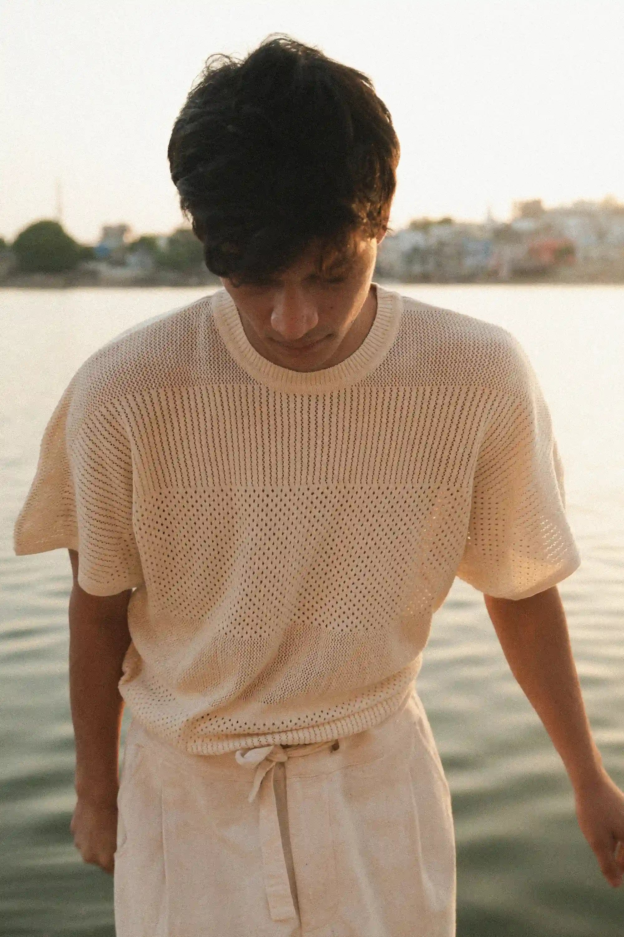 Man wearing a ecru knitted t-shirt and ecru cotton pants by a waterfront.