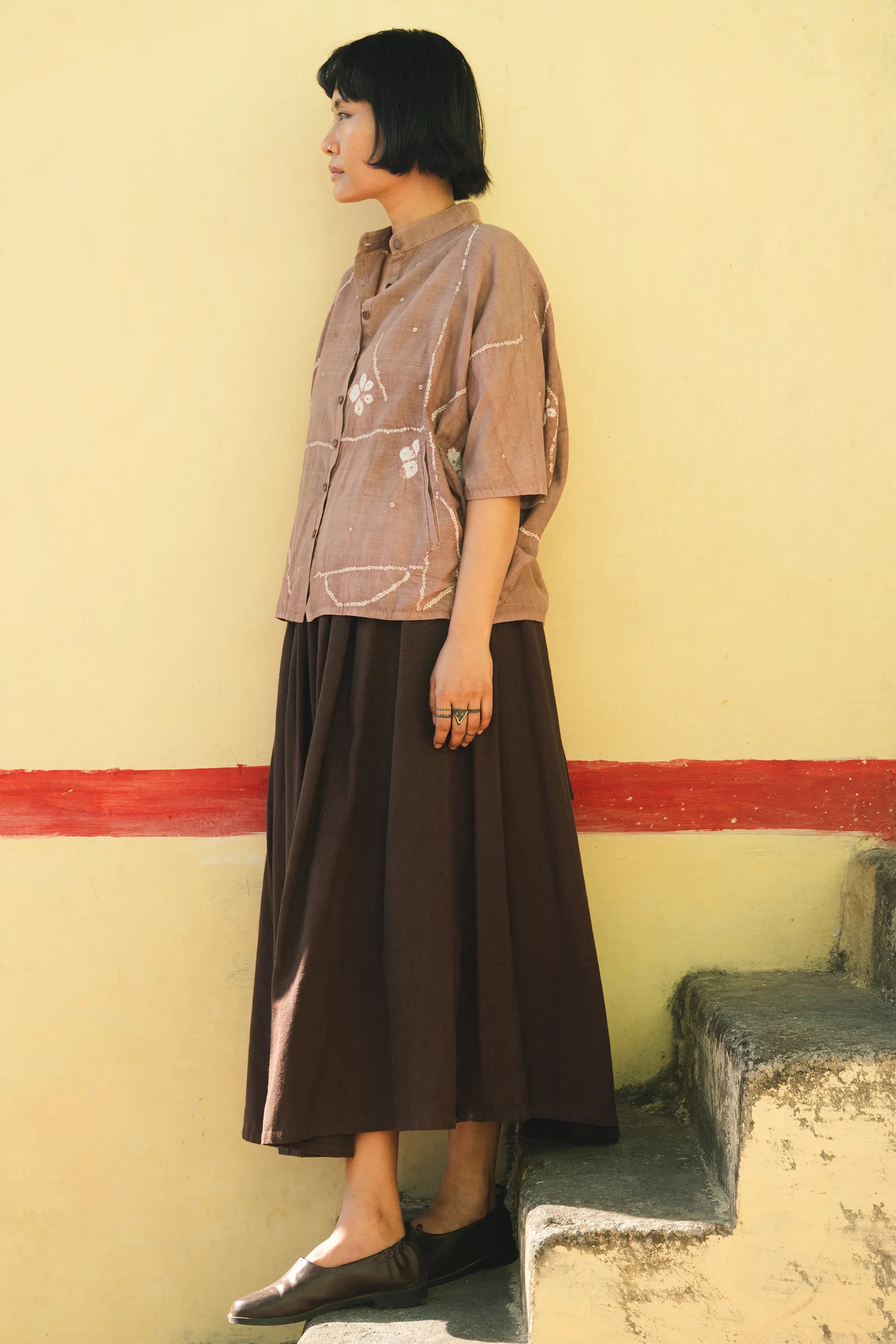 A woman wearing a old rose shirt, burnt umber skirt against a light grey background