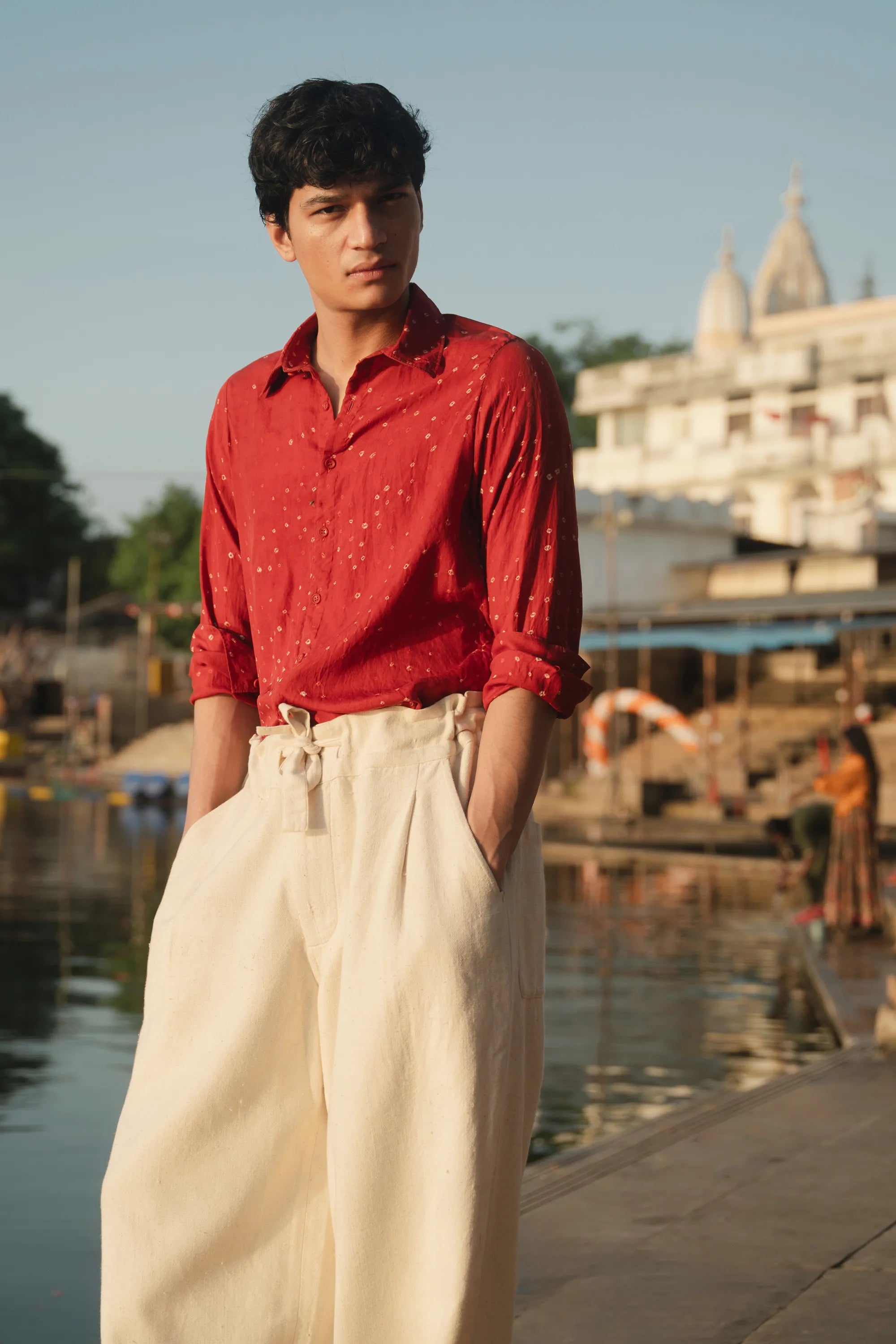 Man in a red shirt and beige pants standing by a body of water with buildings in the background.