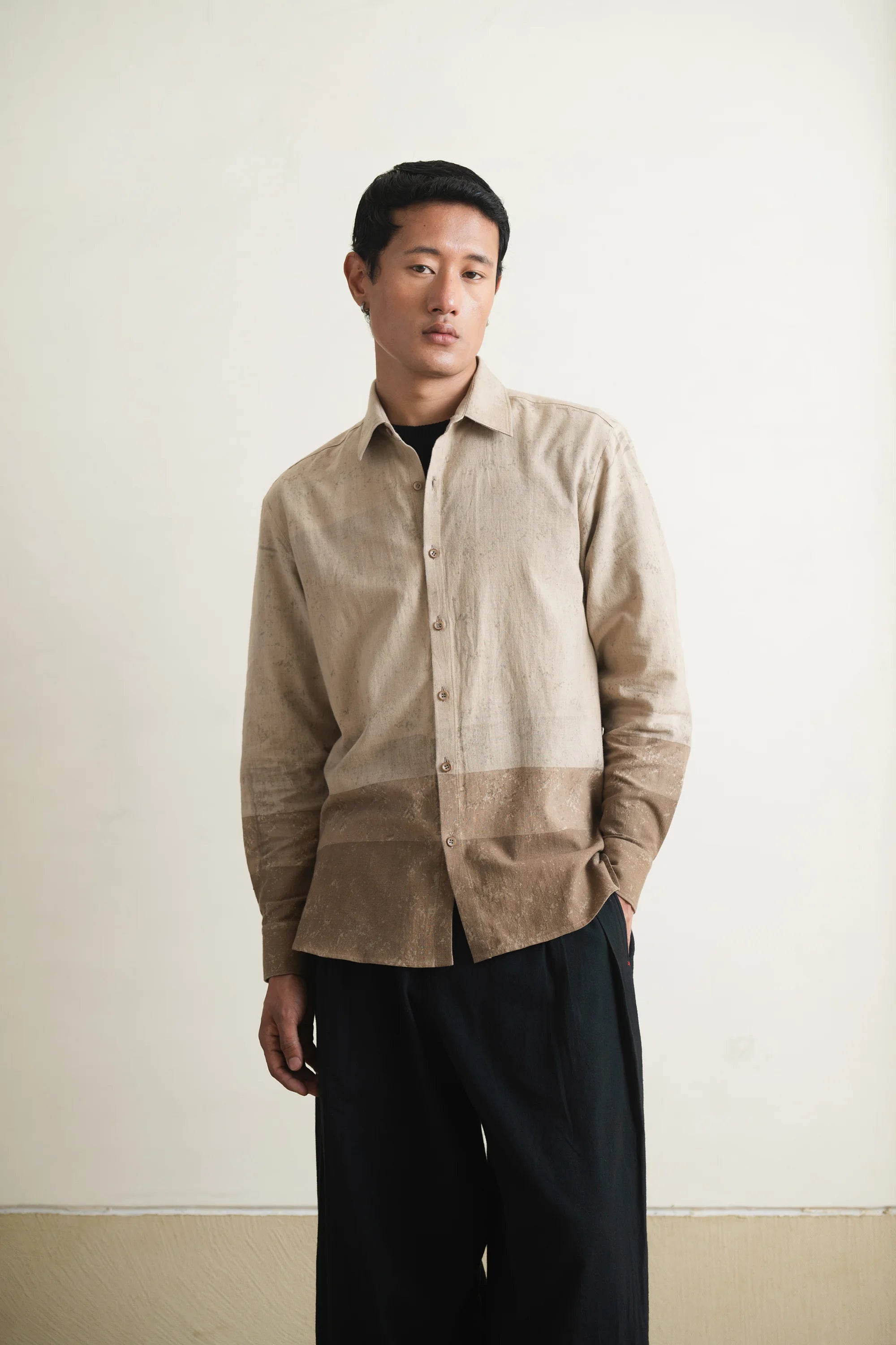 Man wearing a beige and brown shirt with black pants against a plain background