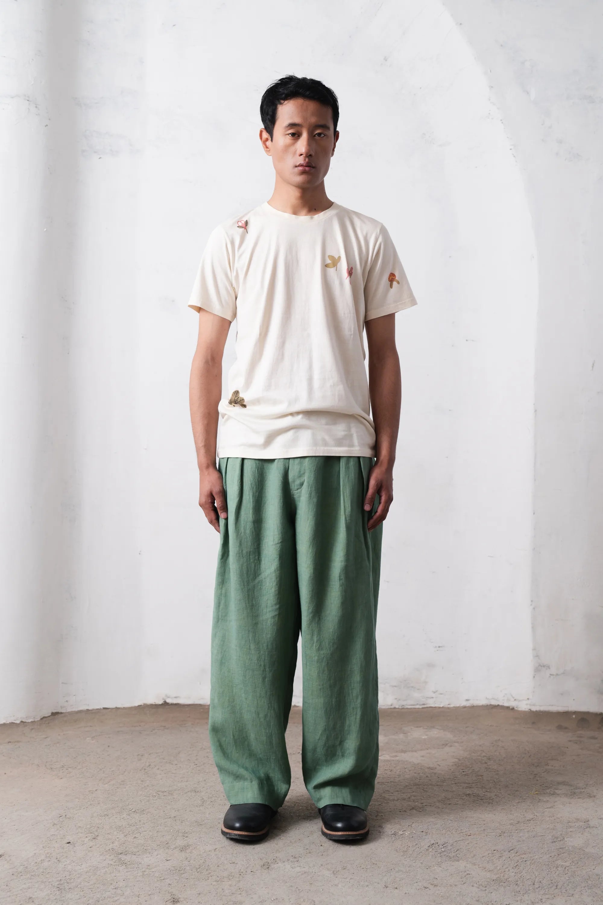 Man wearing a white t-shirt and green pants standing against a light grey wall