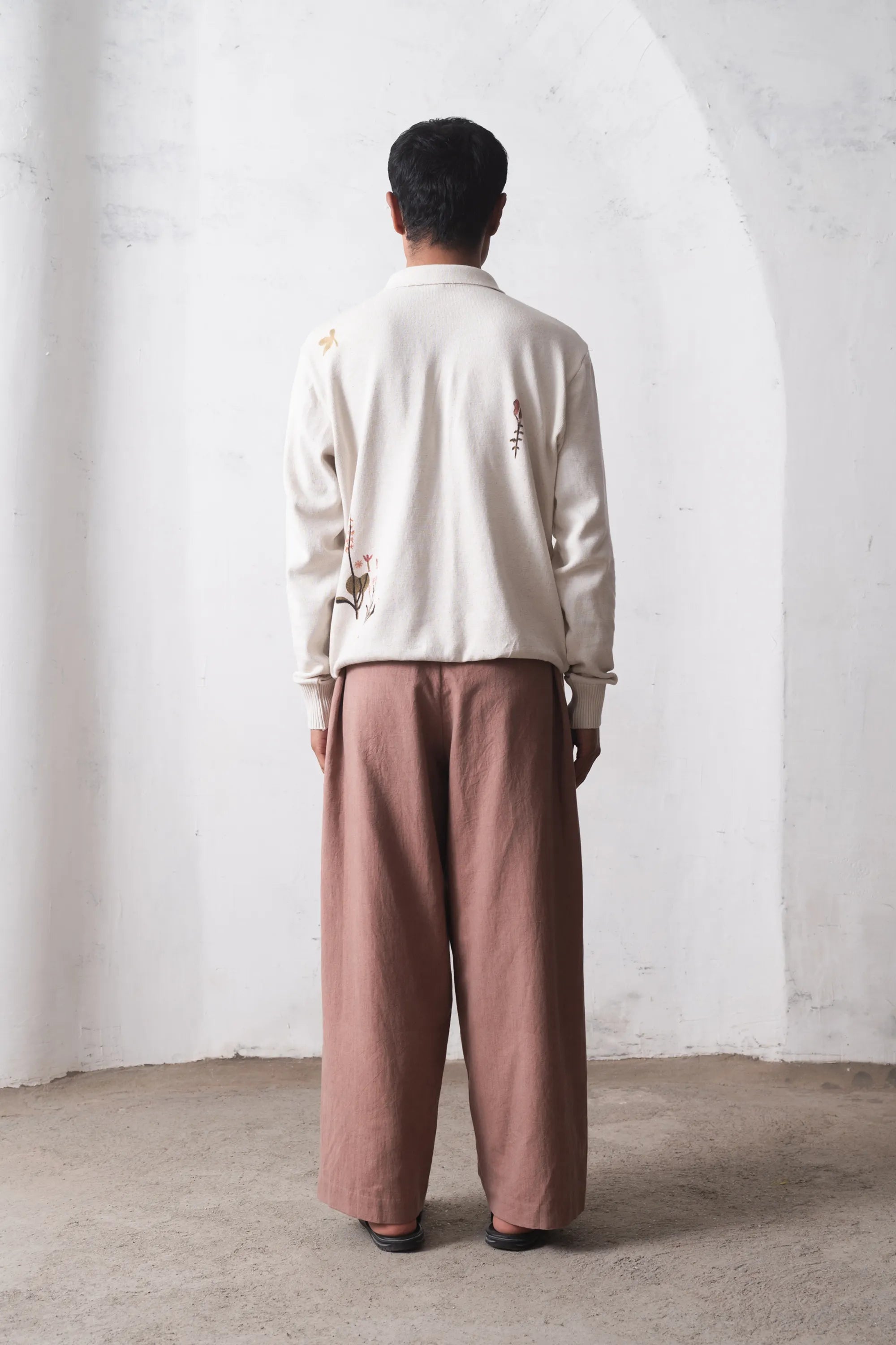 Back shot of a person wearing a ecru t-shirt and brown pants standing against a white wall