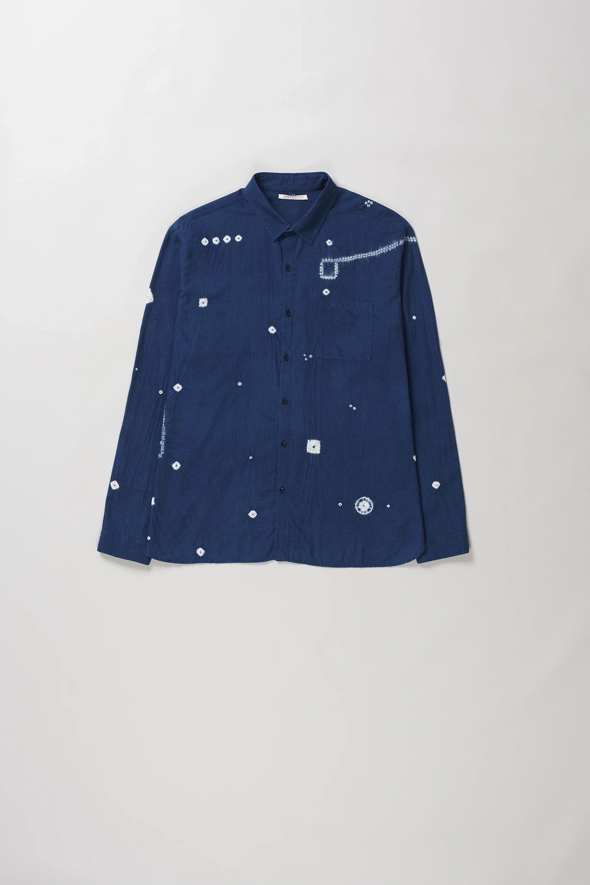 A medium indigo long-sleeve button-up shirt with white shibori details, laid flat against a light grey background.